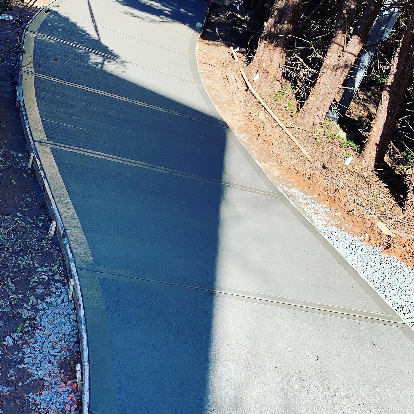 A curved sidewalk under construction next to a dirt and gravel area with trees in the background.
