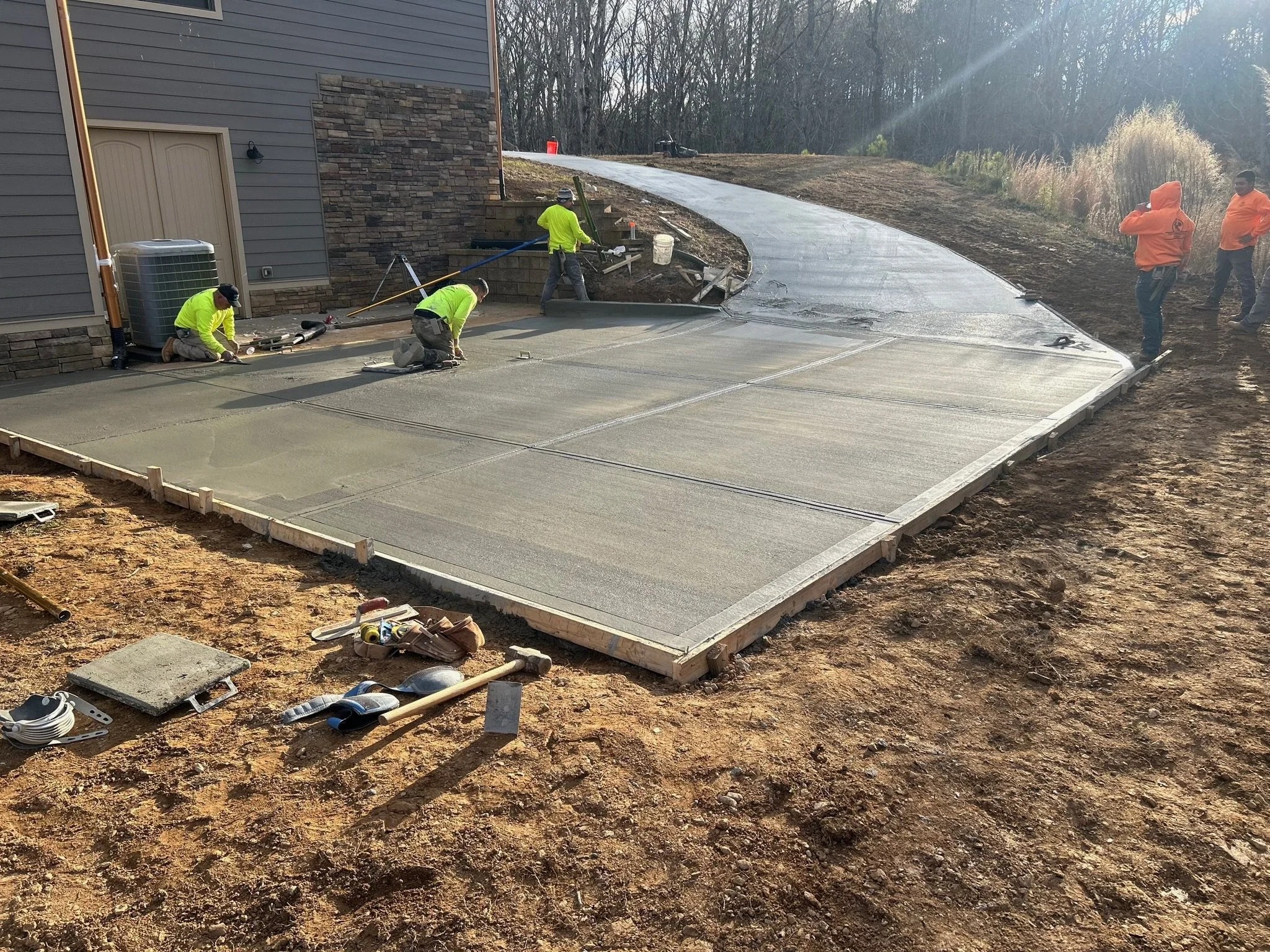 Construction workers laying the foundation for a new concrete driveway, with some workers kneeling and others standing, and building materials scattered on the ground.