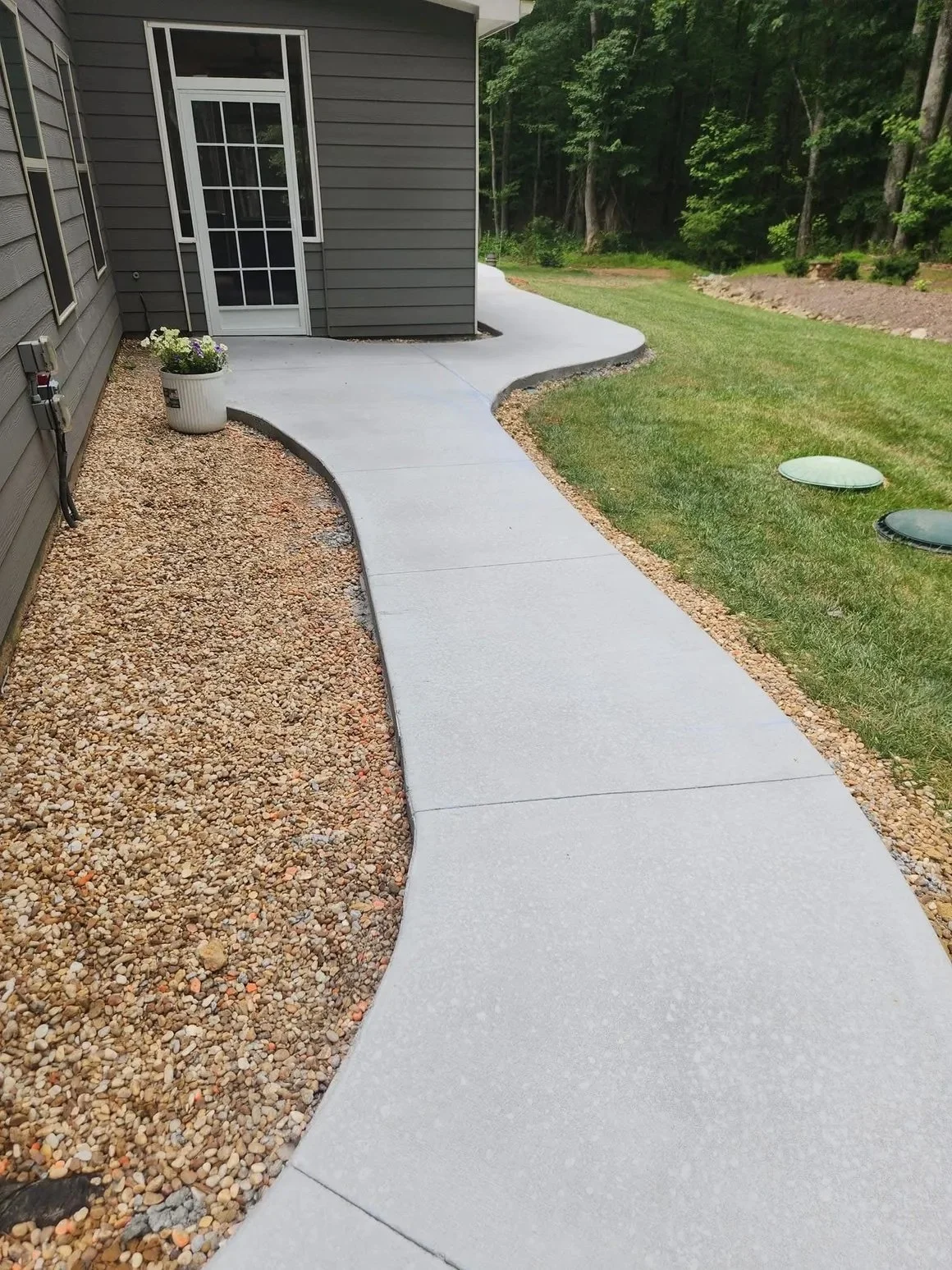 A backyard with a concrete pathway curving around a house, with gravel on one side and a grassy lawn on the other, surrounded by trees.