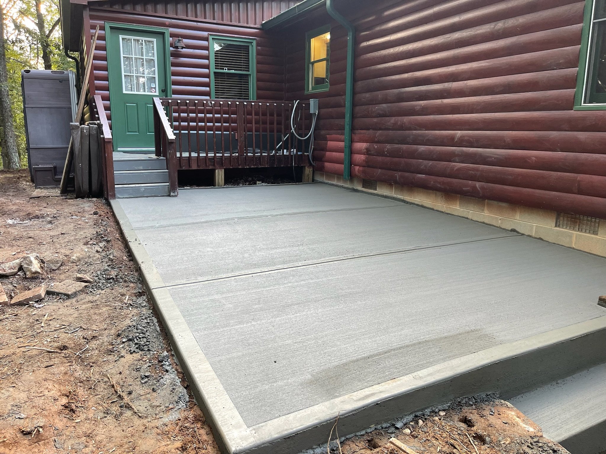 Newly poured concrete patio in front of a wooden house with green trim, with a door, window, and outdoor plumbing fixtures.