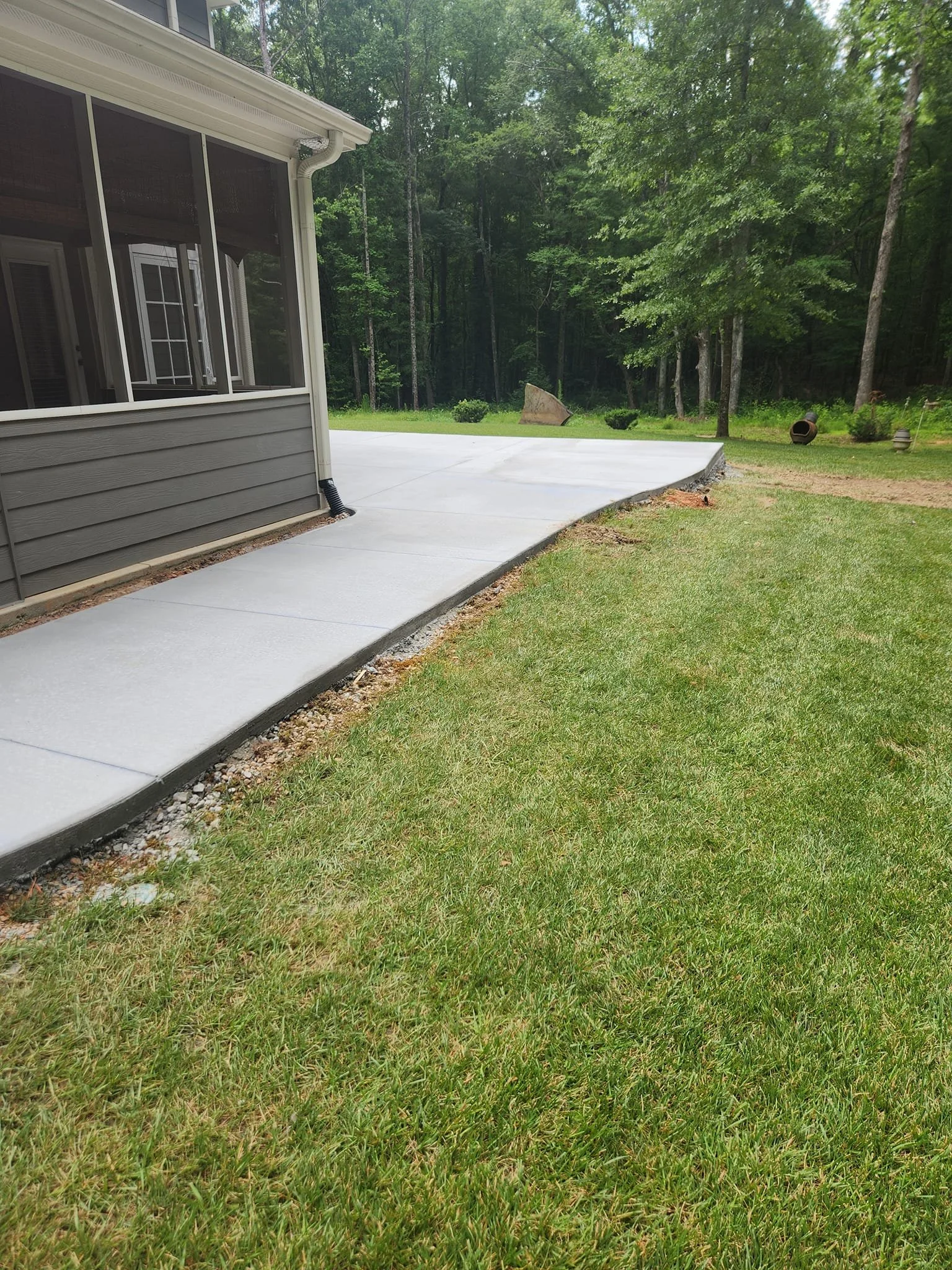 A backyard patio with a concrete slab extending from the house into a green yard surrounded by trees. There are garden ornaments and a barrel near the tree line.