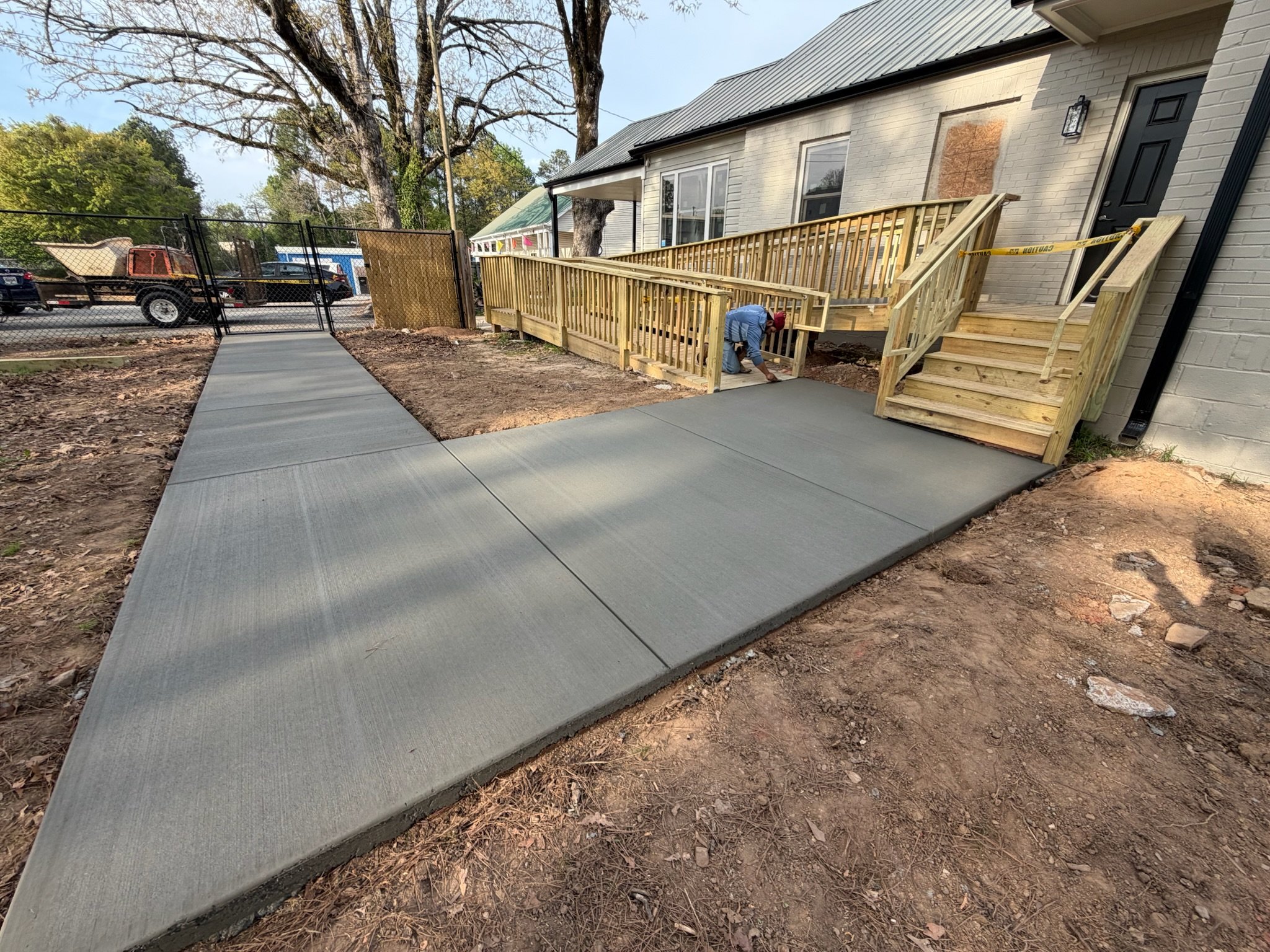 A person installing a wooden ramp and stairs outside a house with a white brick exterior. The area around the construction site has dirt and a newly poured concrete walkway.