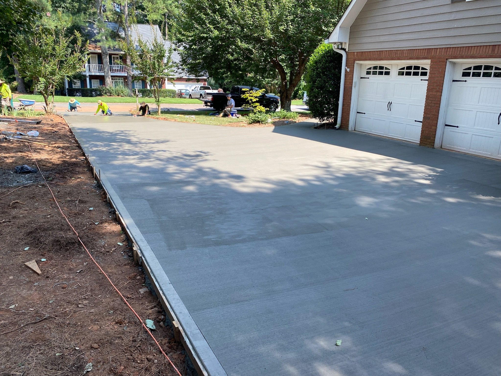 People working on a backyard landscaping project, pouring and smoothing concrete on a driveway, with trees, houses, and parked cars in the background.