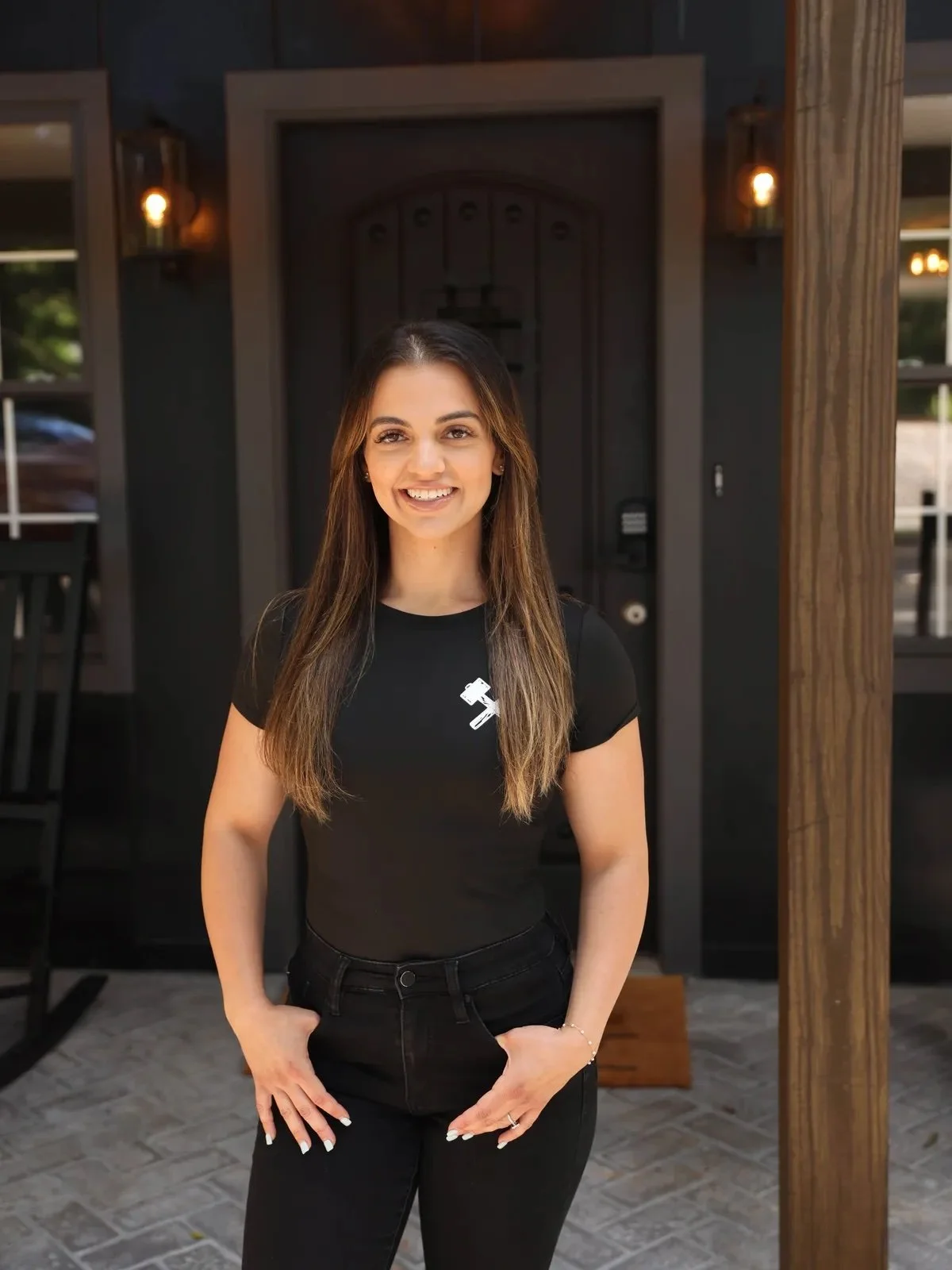 Smiling woman standing in front of a dark background, wearing a black t-shirt with a white logo and black pants.
