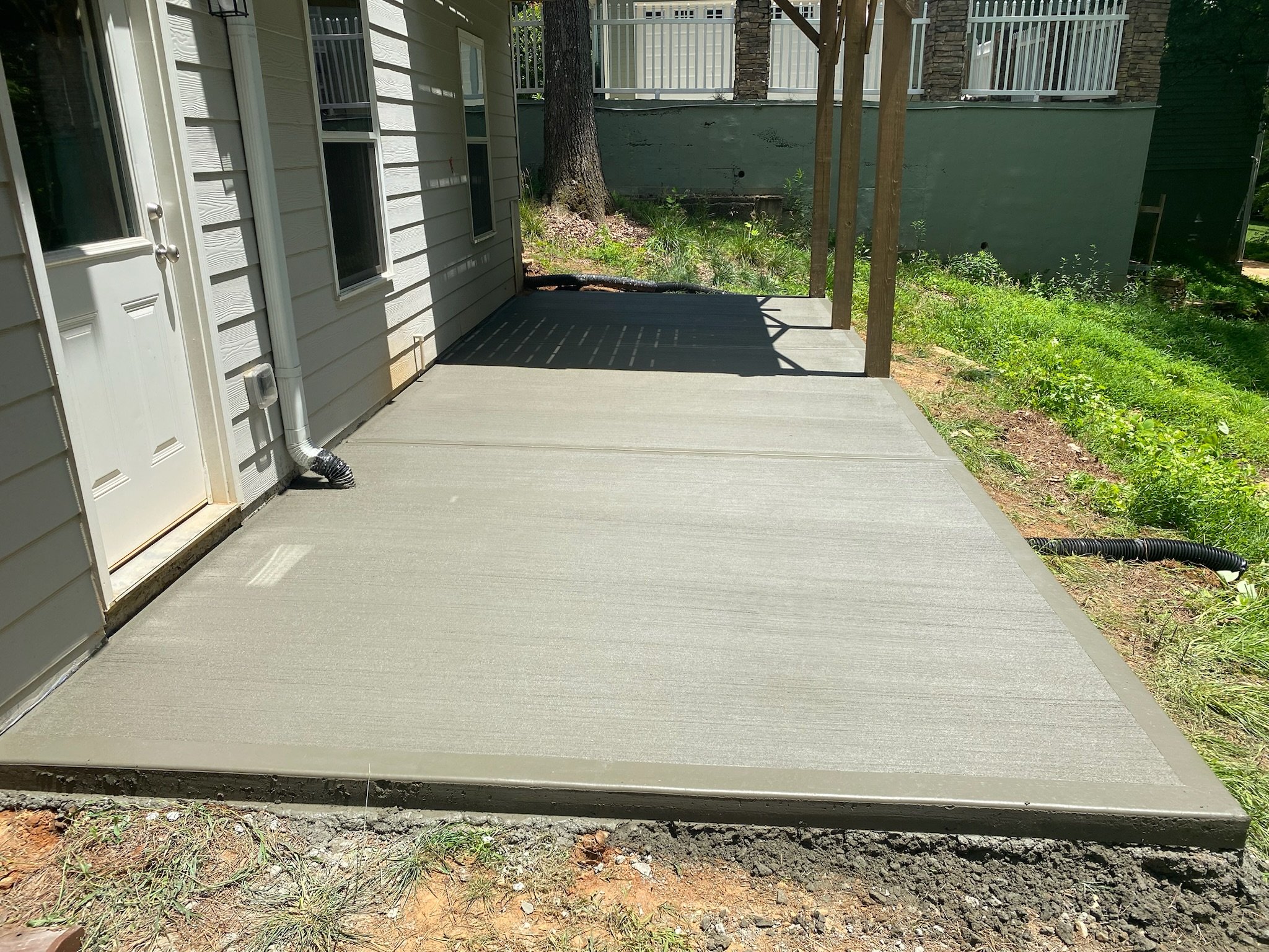 A newly poured concrete patio with a smooth surface, attached to a house with white siding, and a small grassy area with a tree and some plants behind the patio. The house has windows and a door, and the patio is framed with wooden posts for a future