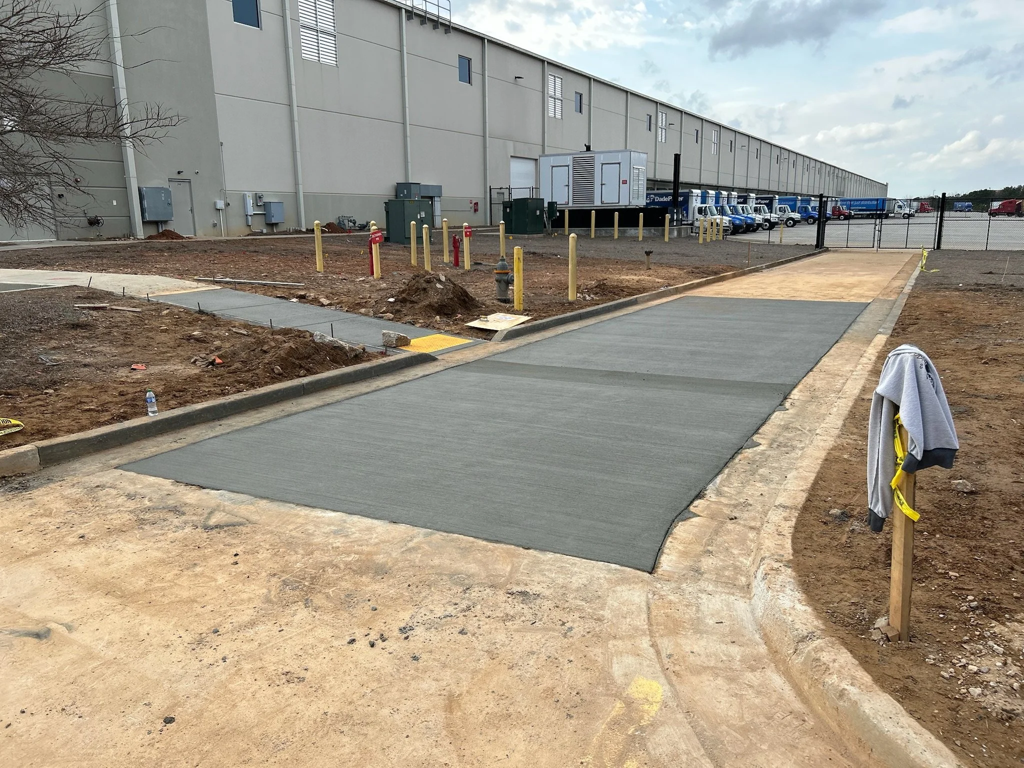 Construction site with unfinished sidewalk, dirt, and construction materials, next to a large industrial building, with a parking lot of trucks and vehicles in the background.