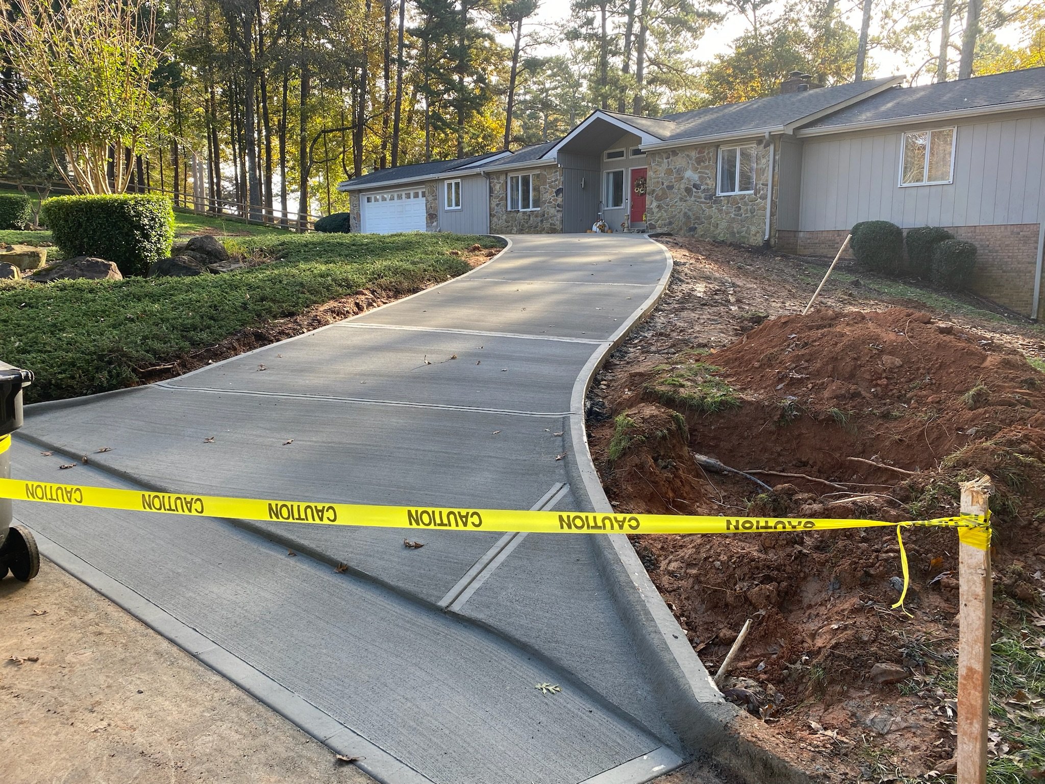 A newly paved driveway leading up to a house with a stone and gray exterior, surrounded by a wooded yard. Yellow caution tape is placed at the edge of the construction zone.