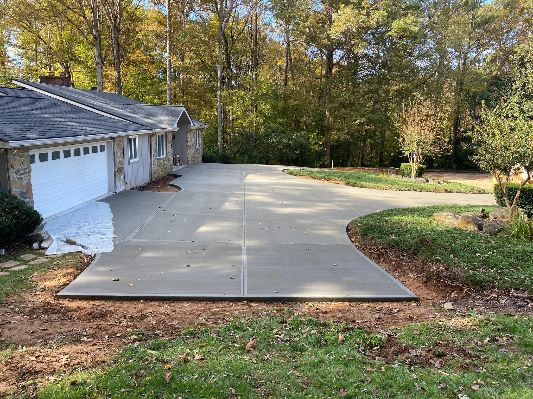 A newly paved concrete driveway with a gentle curve leading to a garage, surrounded by a garden with trees and shrubs, and a wooded area in the background.