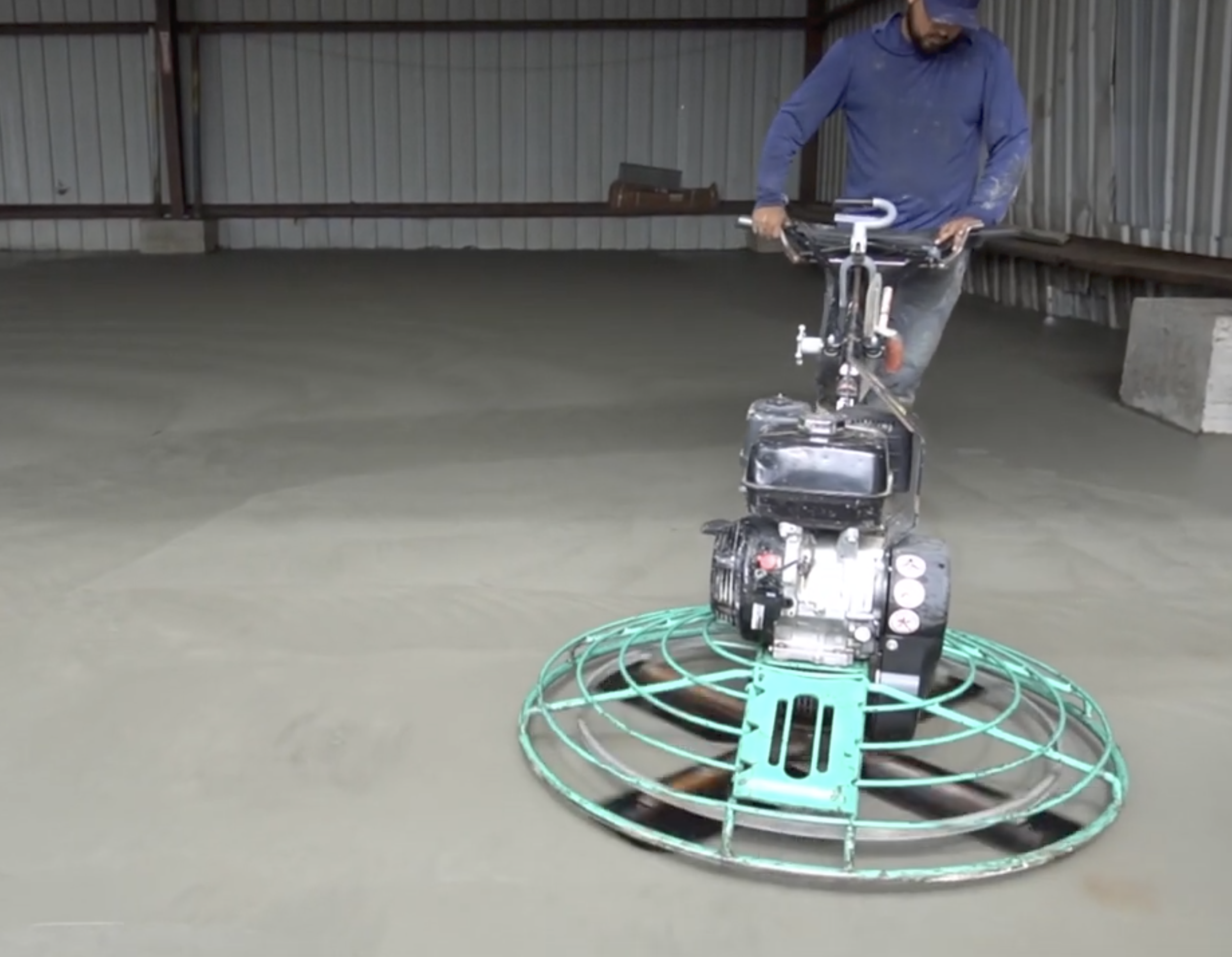 A person standing in an indoor space, possibly a warehouse or workshop, with a BMX bike and a coiled green cable or hose on the floor. The person is wearing a blue long-sleeve shirt and a cap.