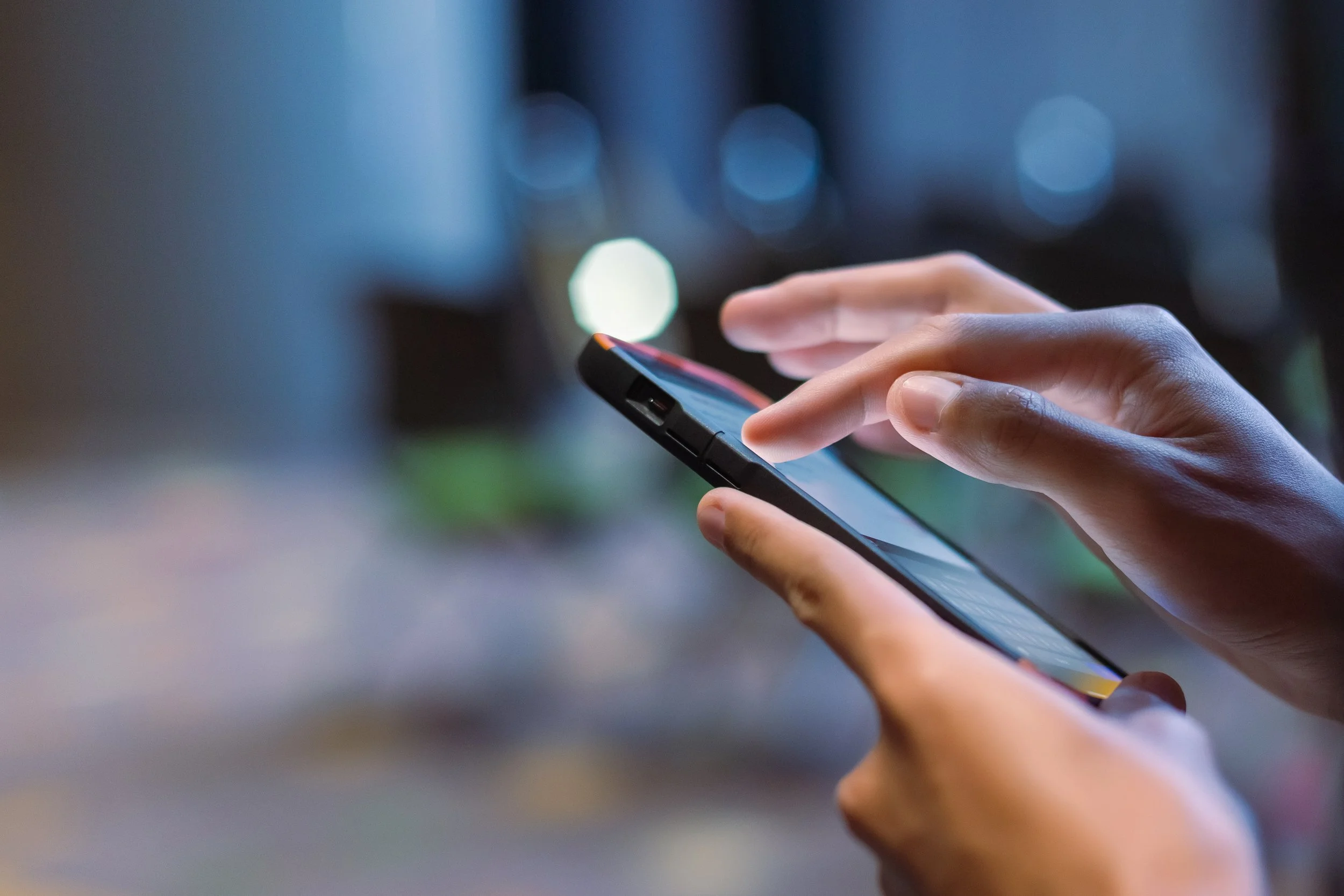 Close-up of a person using a smartphone at night with blurred city lights in the background.