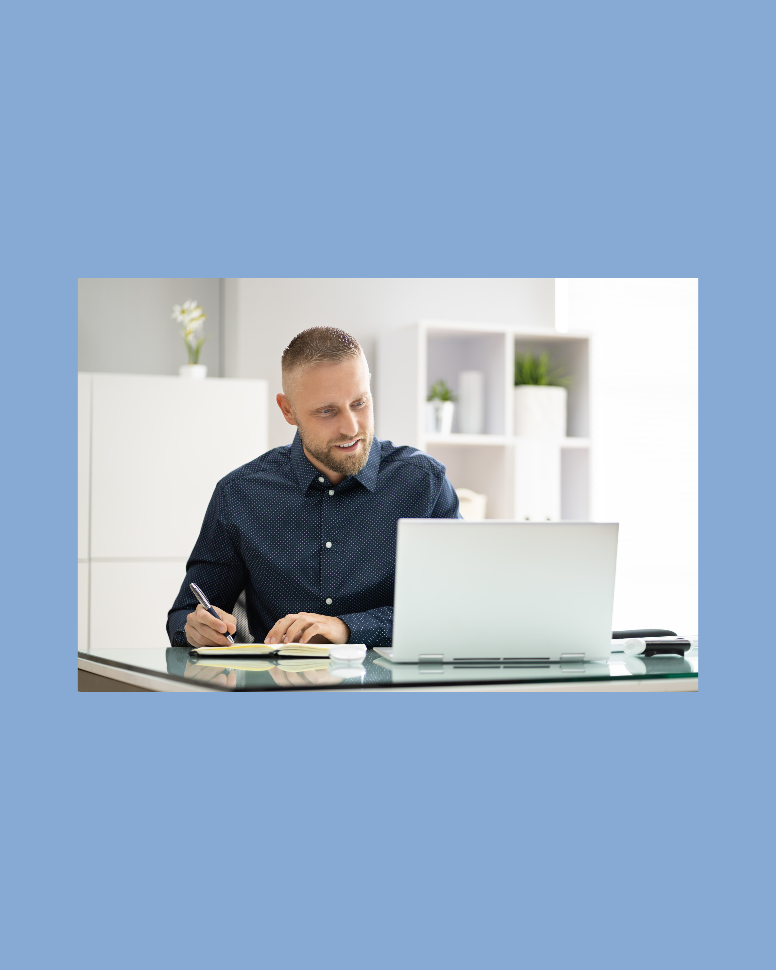 A man with short hair and a beard, wearing a dark blue patterned shirt, working at a desk. He is writing in a notebook with a pen. There is a laptop open in front of him, and the background shows white shelves with decorative items and plants.