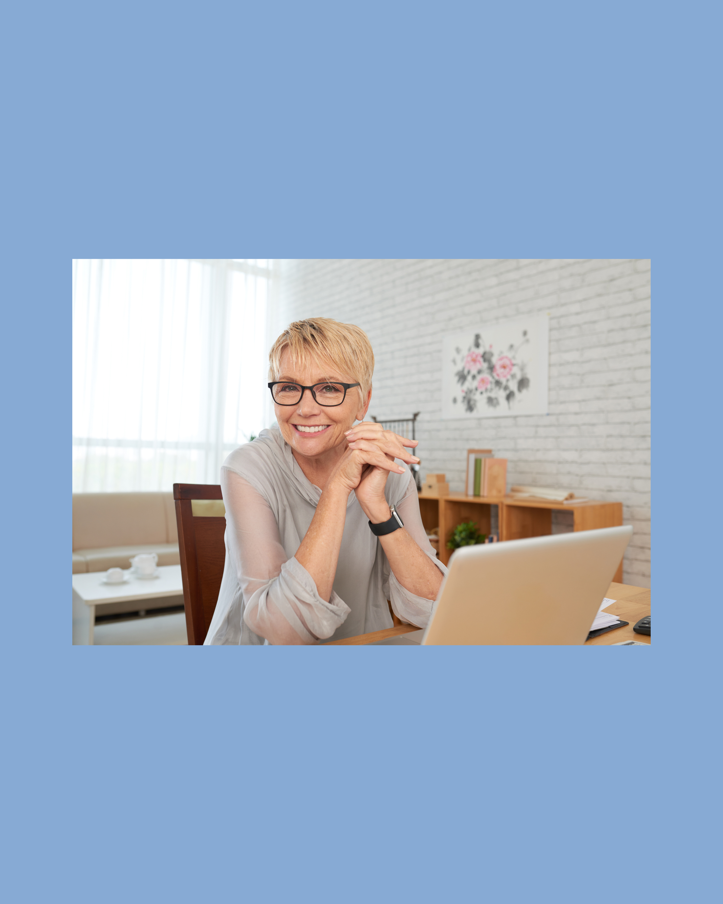 A woman with short blonde hair, glasses, and a light gray shirt sitting at a wooden desk with a laptop, smiling, in a bright room with large windows, white brick wall, and a floral painting in the background.