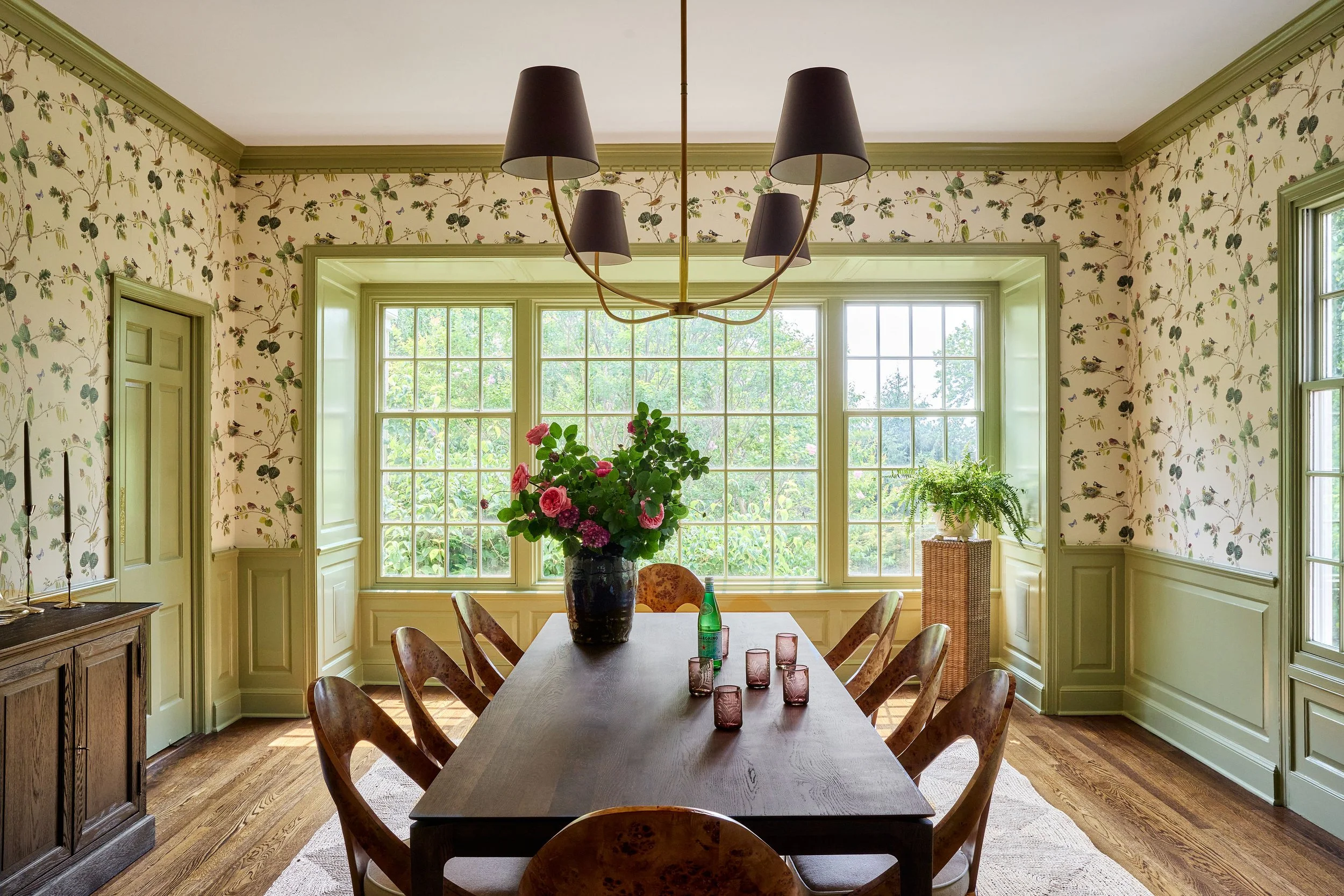 A dining room with a wooden table and chairs, large windows with green trim, floral wallpaper, and a chandelier with black lampshades.