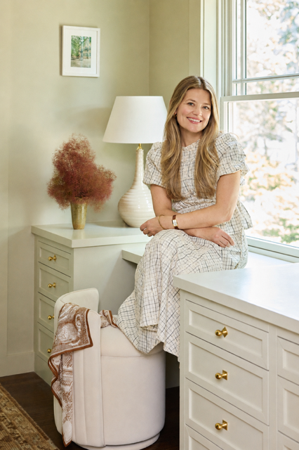 A woman in a checkered dress sitting on a white armchair next to a window in a bright room. There are white drawers, a table lamp, a vase with dried flowers, and a framed picture on the wall.