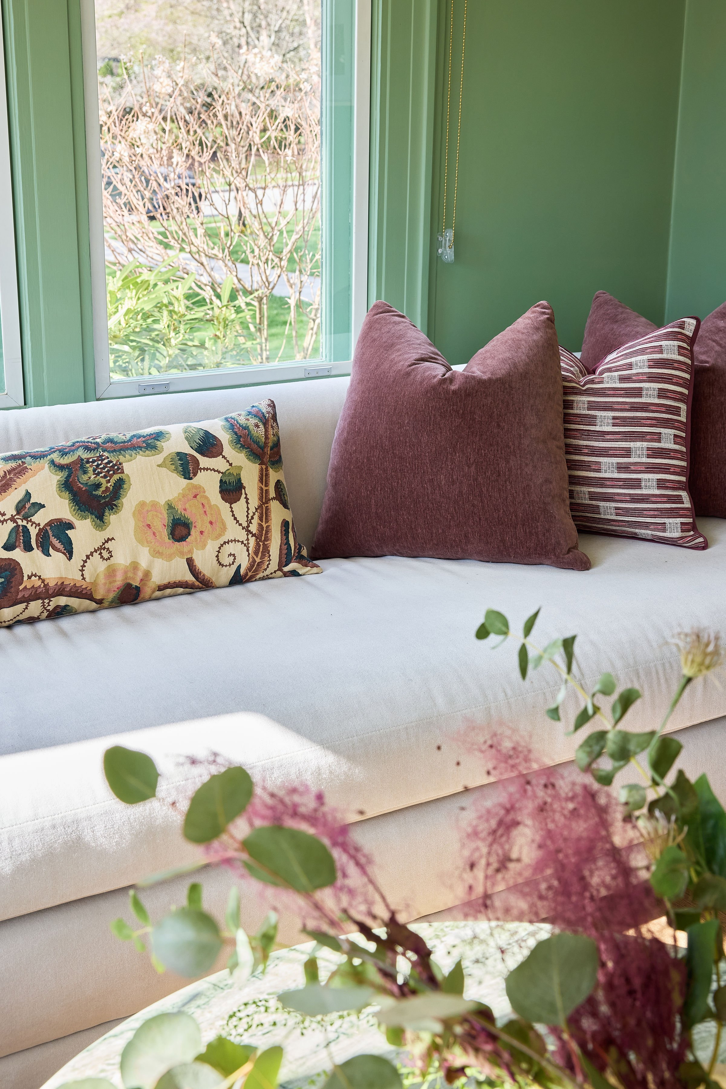 A cozy indoor space with a white sofa decorated with patterned and solid-colored pillows, overlooking a garden through large green-framed windows.