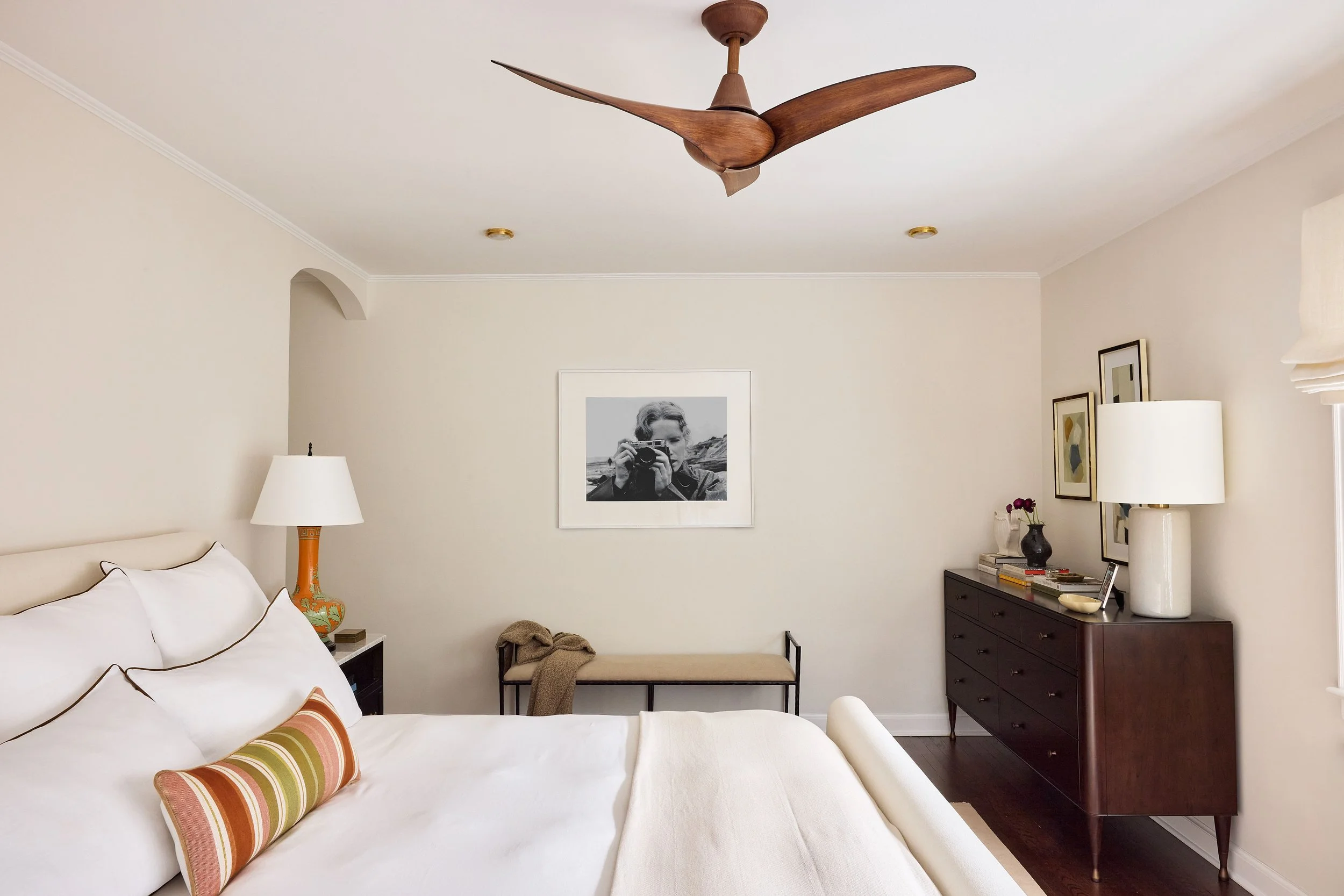 Interior of a bedroom with a white bed, colorful throw pillows, bedside lamp, black-and-white photograph on the wall, black dresser, and decorative items.