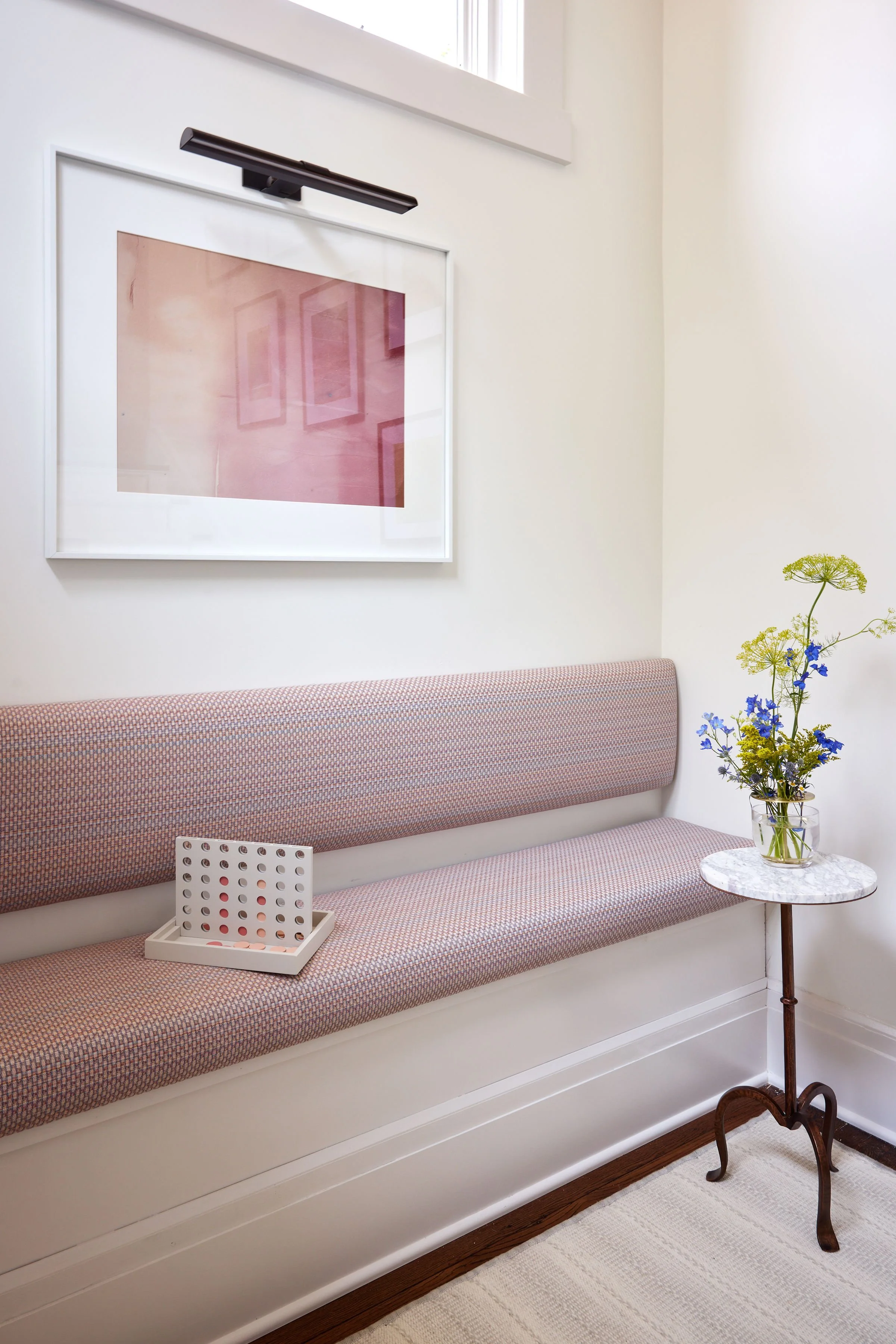 A waiting room with a pink upholstered bench, a small round marble-topped side table holding a glass vase with yellow and blue flowers, a framed pink artwork on the wall, a white wall and a window at the top, a white plastic tray with a Connect 4 game on the bench, and a beige and white striped rug.