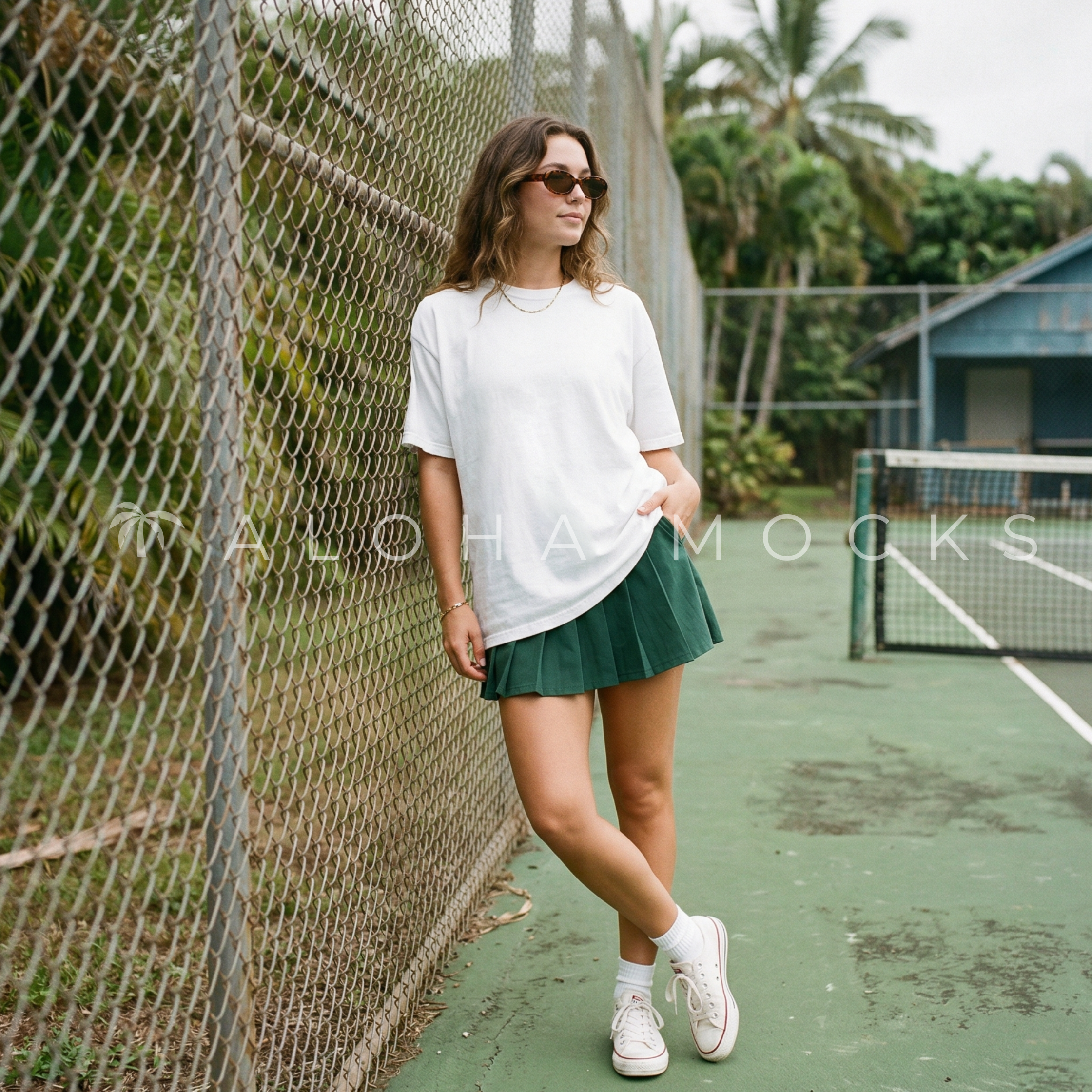 A woman in sunglasses and a white t-shirt with a green skirt leaning against a chain-link fence on a tennis court with trees in the background.