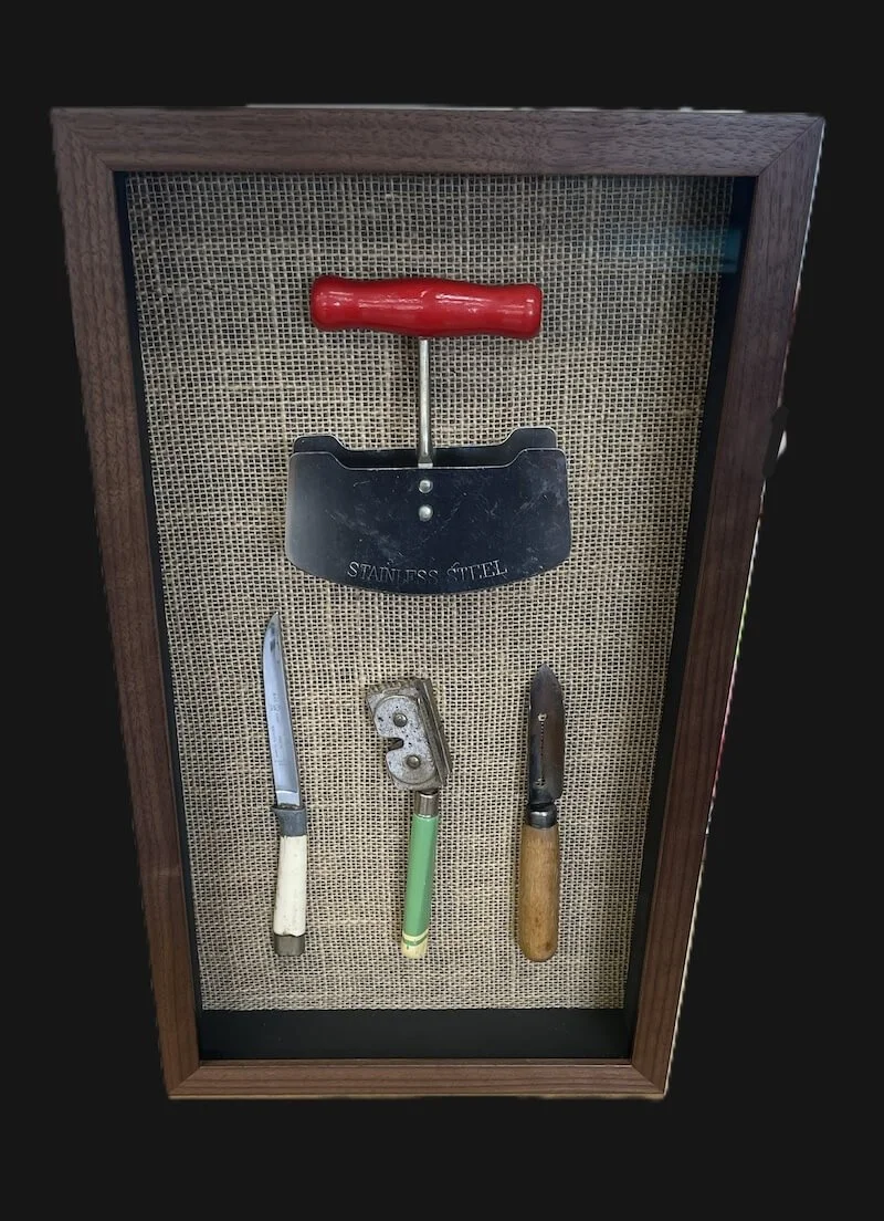 A display case holding four vintage tools: a small knife with a white handle, a vintage can opener with a green handle, a small hand axe with a wooden handle, and a steel spatula with a red handle.