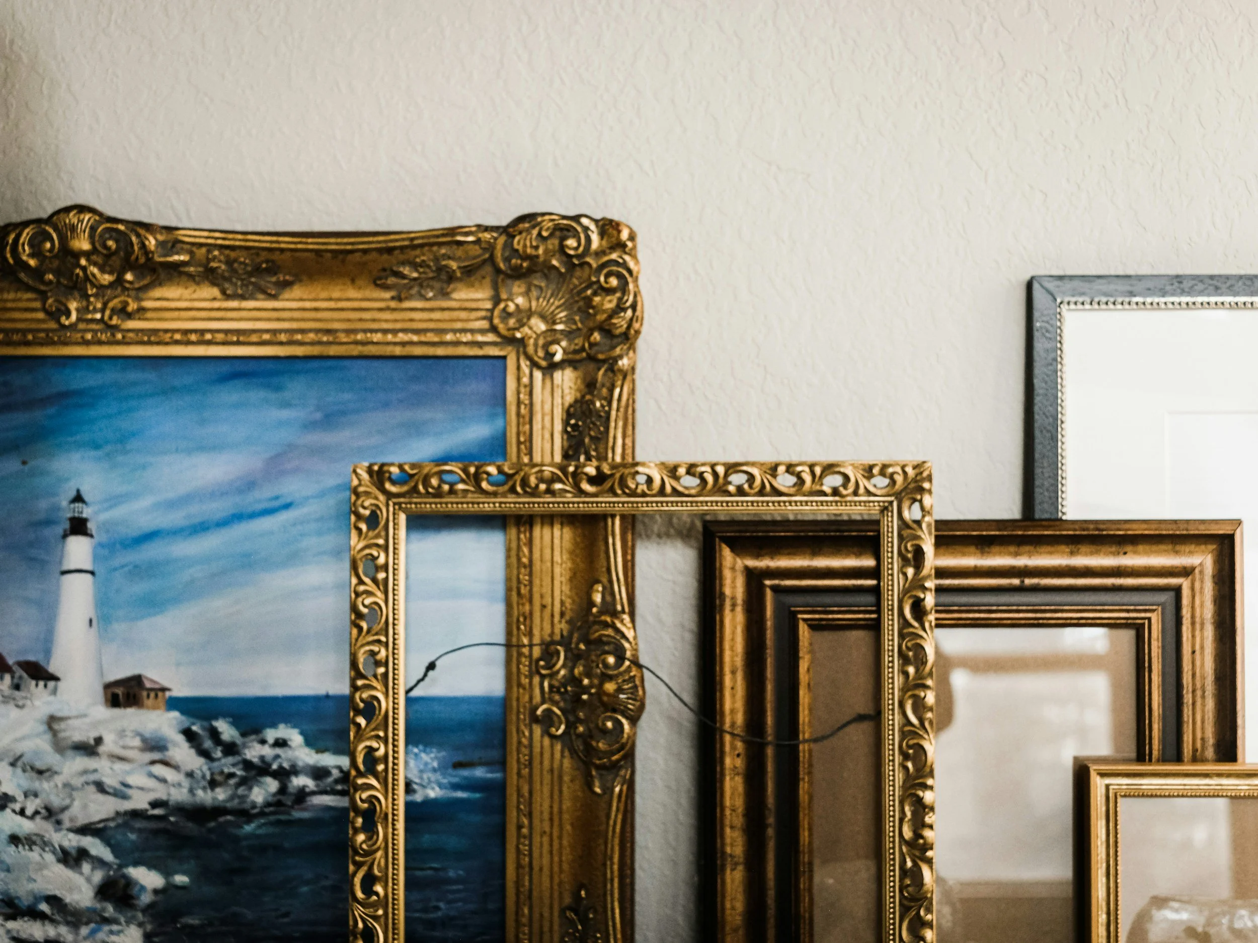 Multiple empty decorative picture frames are stacked against a textured white wall in a room. A painting of a lighthouse on rocky coast is partially visible behind the frames.