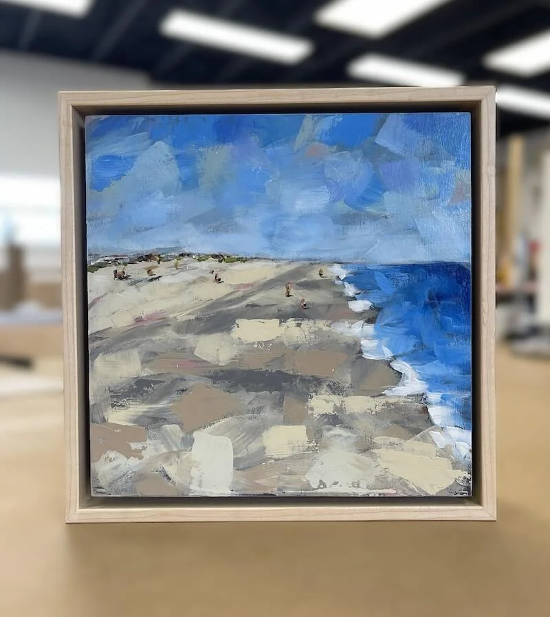 A small impressionist-style painting of a beach with sand, ocean waves, and a cloudy sky, framed in light wood.