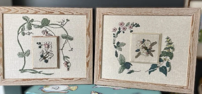 Two embroidered floral pictures with wooden frames, each featuring a central flower surrounded by vine and leaf patterns.