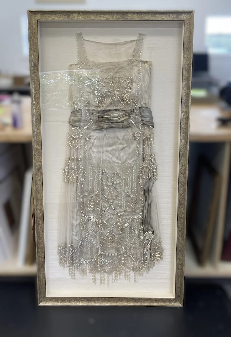 Framed and preserved vintage dress with intricate beadwork and lace details, displayed in a shadow box.