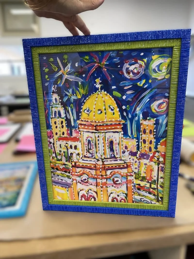 Colorful painting depicting a cityscape with a large domed building and fireworks in the night sky, framed in a blue and green frame.
