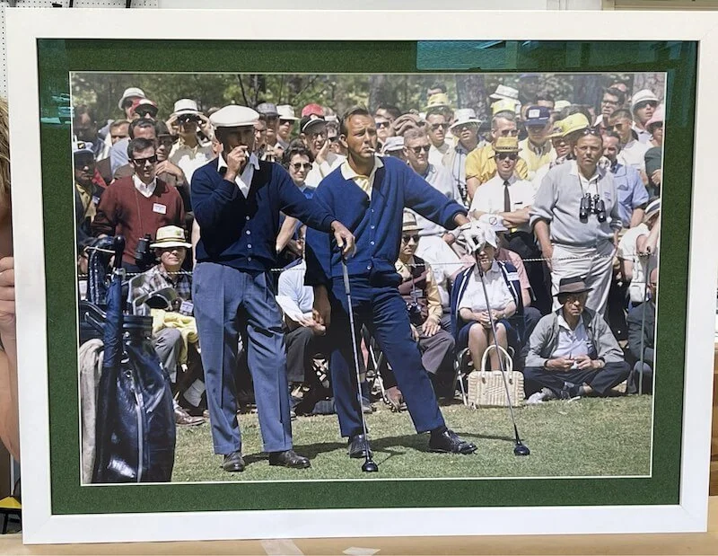 Vintage golf scene with two men in navy sweaters on a golf course, surrounded by a large crowd of spectators wearing hats and 1950s attire.