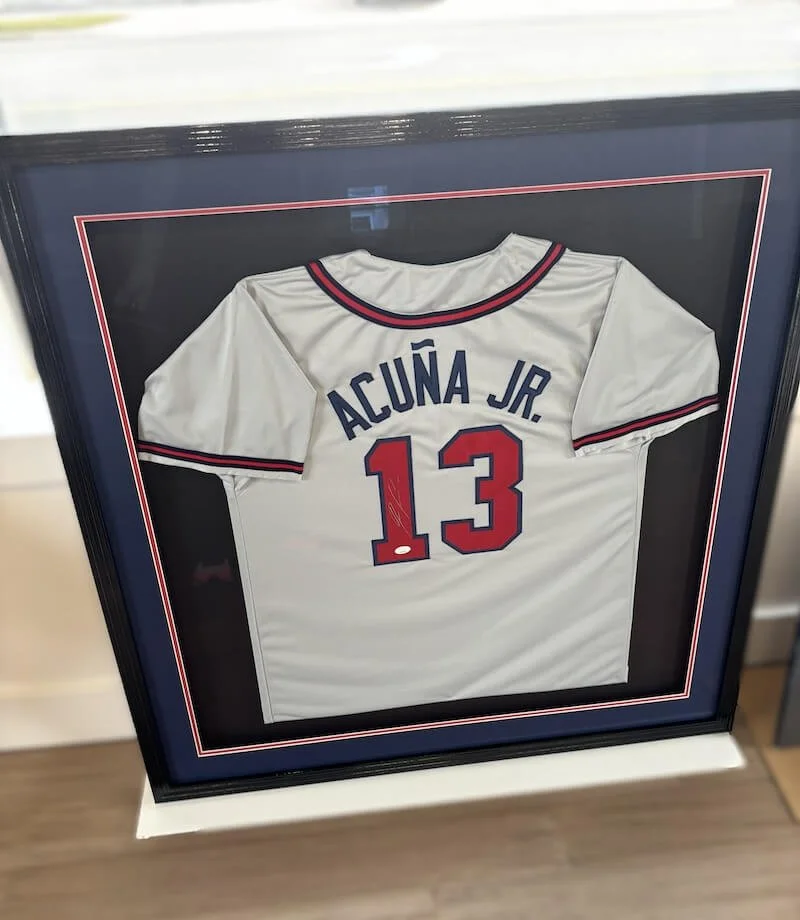 Framed white baseball jersey with red and navy accents, displaying the name 'Alcúna Jr.' and the number 13.