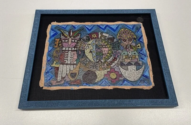 A framed textile artwork depicting two stylized cartoon-like characters, one holding a trumpet and the other a staff, with geometric patterns and symbols in the background.