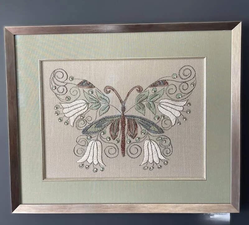 Framed embroidered artwork of a butterfly with swirled antennae and floral design on its wings.