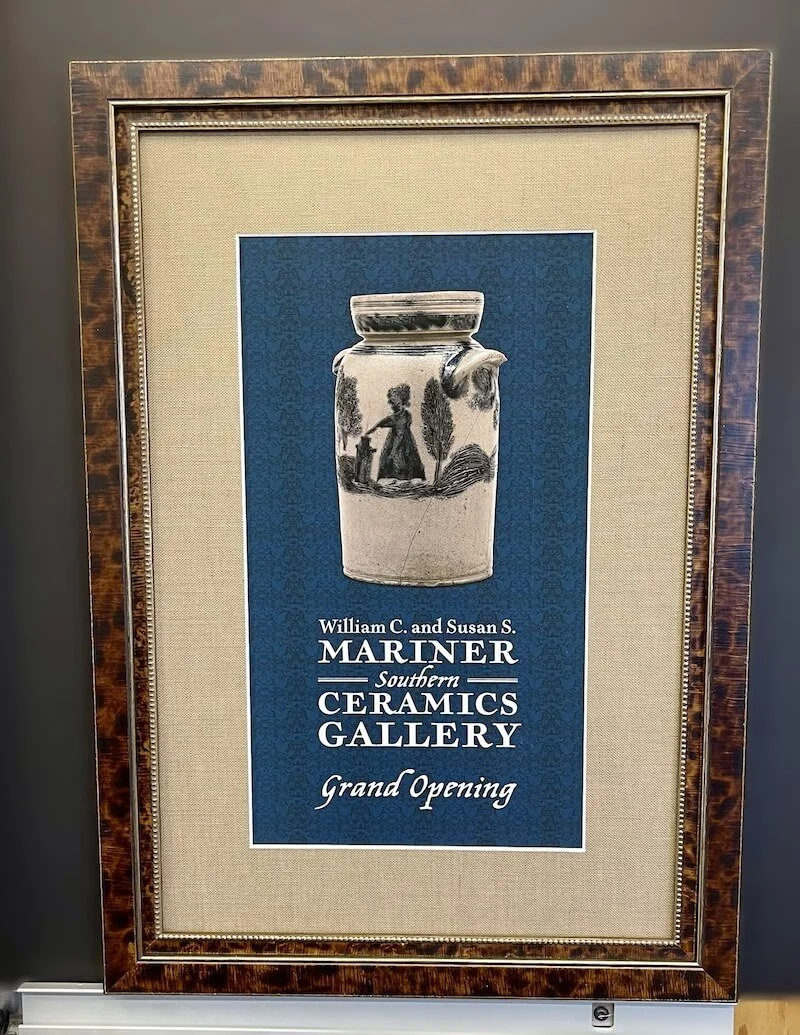 Framed announcement for William C. and Susan S. Mariner Southern Ceramics Gallery Grand Opening, featuring a black-and-white picture of a ceramic jar with painted figures and scenery in a dark blue background.