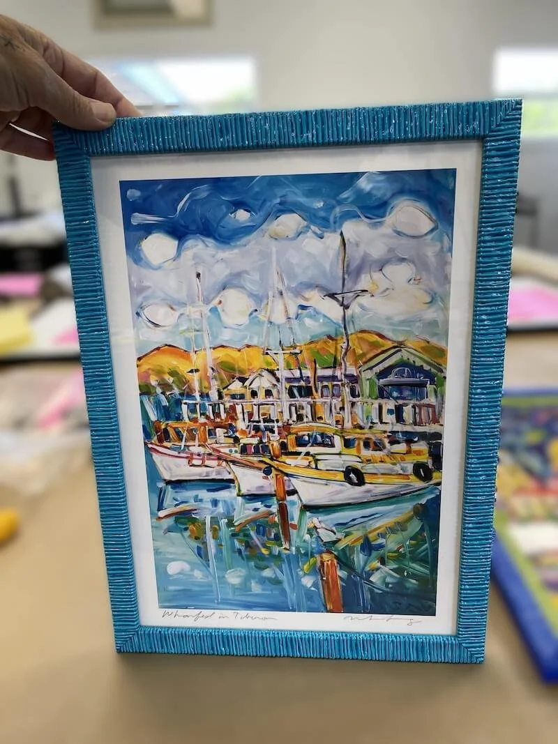 Colorful watercolor painting of a lakeside scene with boats, docks, a house, and hills in the background, in a blue textured frame.