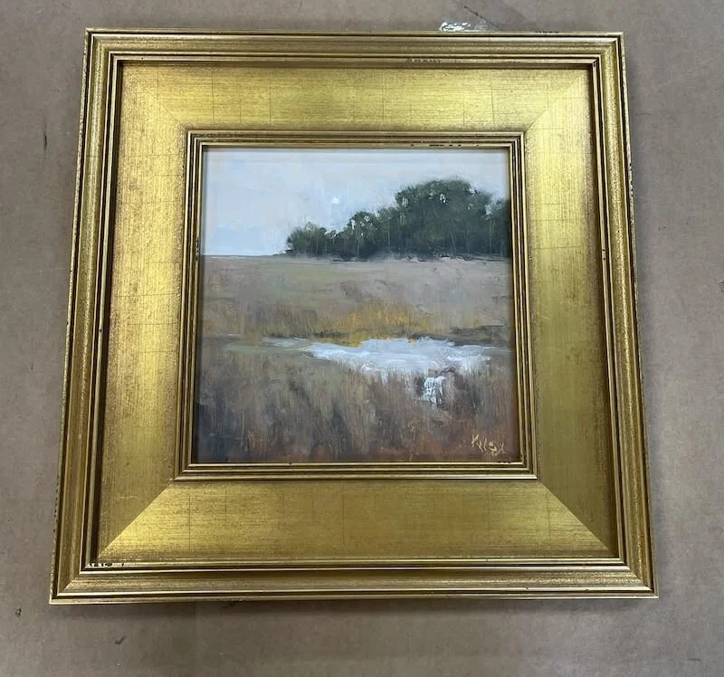 Small landscape painting of a river flowing through a grassy field with trees in the background, framed with a gold ornate frame.