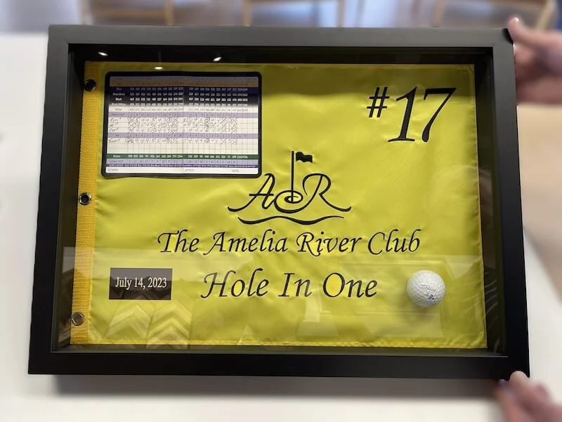 A framed award or certificate for the 'Hole In One' achievement at The Amelia River Club, dated July 14, 2023, featuring a golf ball, a small scoreboard, and decorative text.