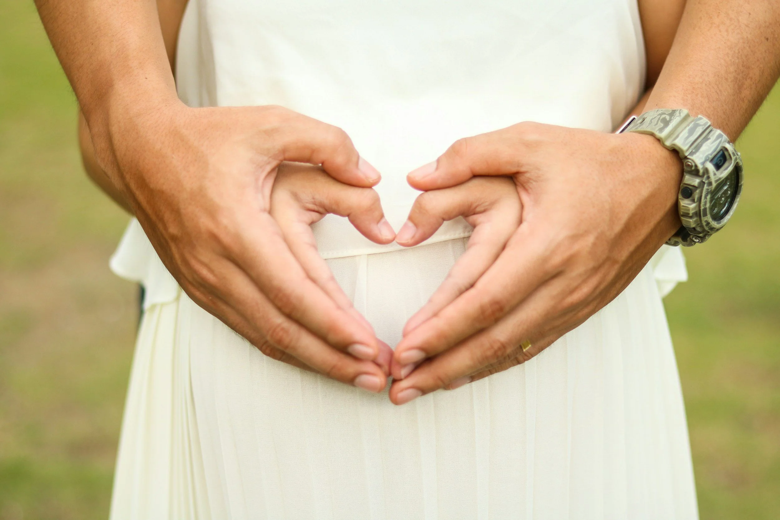 Person making a heart shape with hands over their stomach, wearing a beige watch on wrist, outdoors with blurred green background.