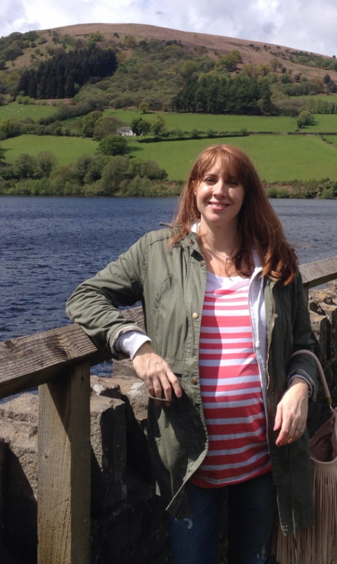 A pregnant woman with red hair smiling, standing in front of a lake with green hills and a mountain in the background.