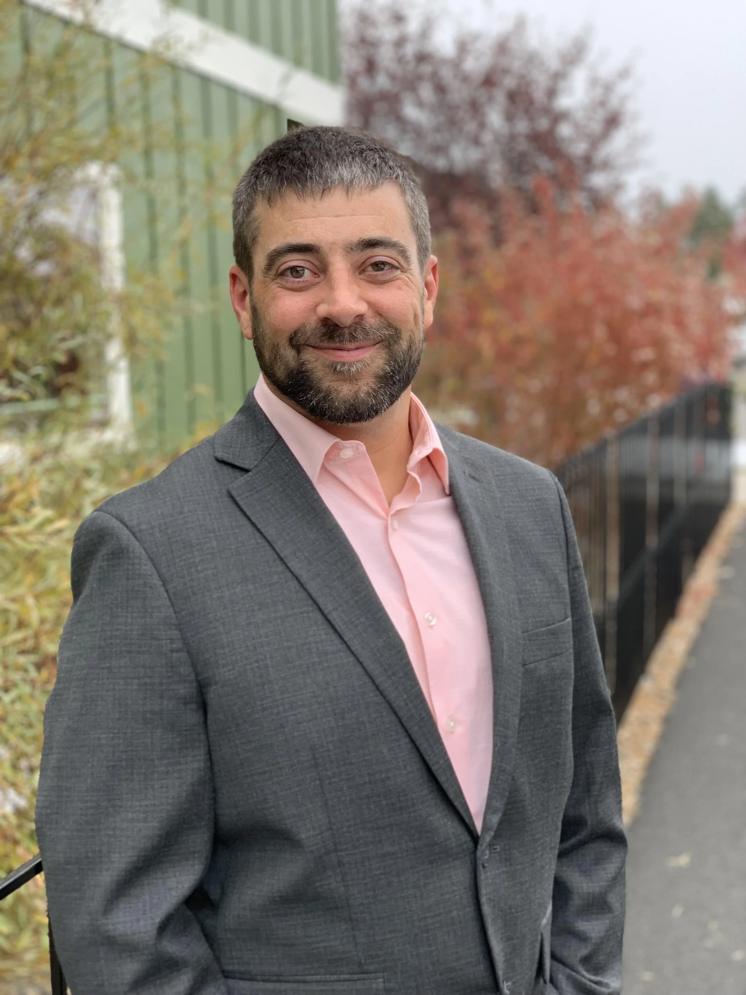 Headshot of a smiling man with short dark hair, beard, wearing a grey suit and pink shirt, outdoors with trees and a green fence in the background.
