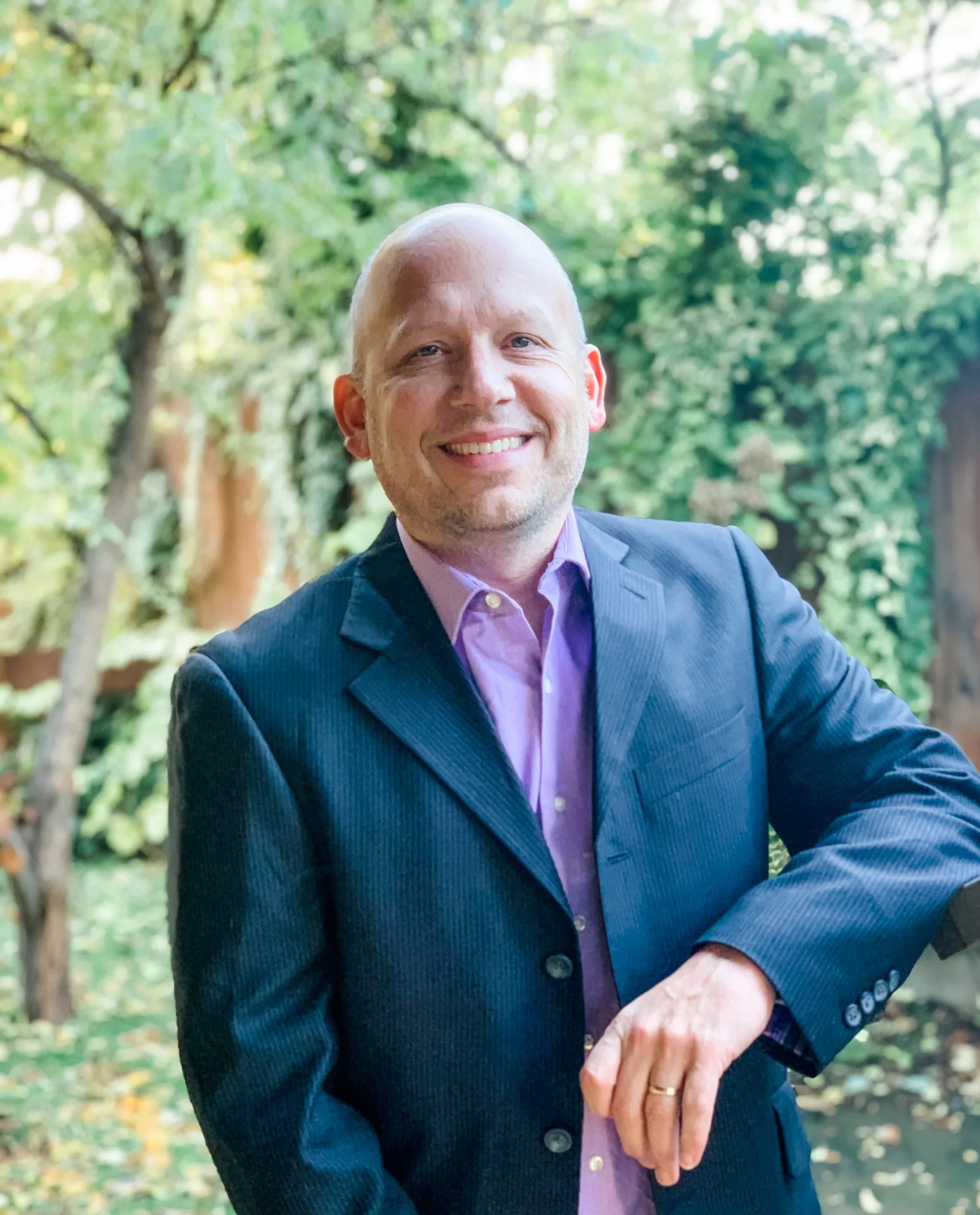A smiling man in a navy suit and light purple shirt shaking hands outdoors with a green leafy background.
