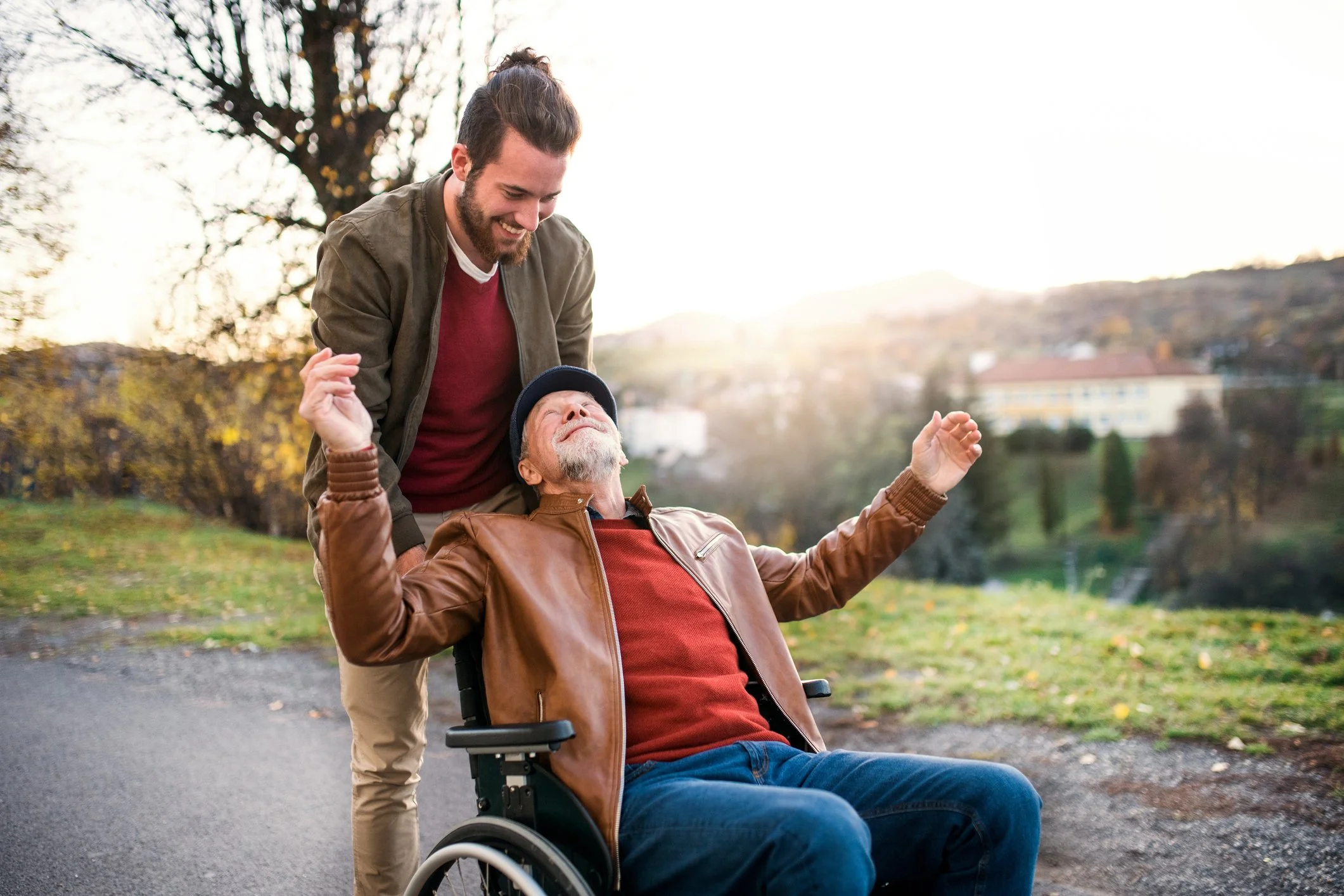 A man with a beard and a woman with gray hair riding in a wheelchair outdoors in a park. The woman is smiling with her arms raised, and the man is standing behind her, smiling down at her. Trees and a hillside are visible in the background.