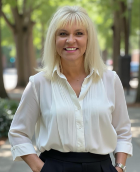 A smiling woman with blonde hair wearing a white blouse standing outdoors in a park with green trees.