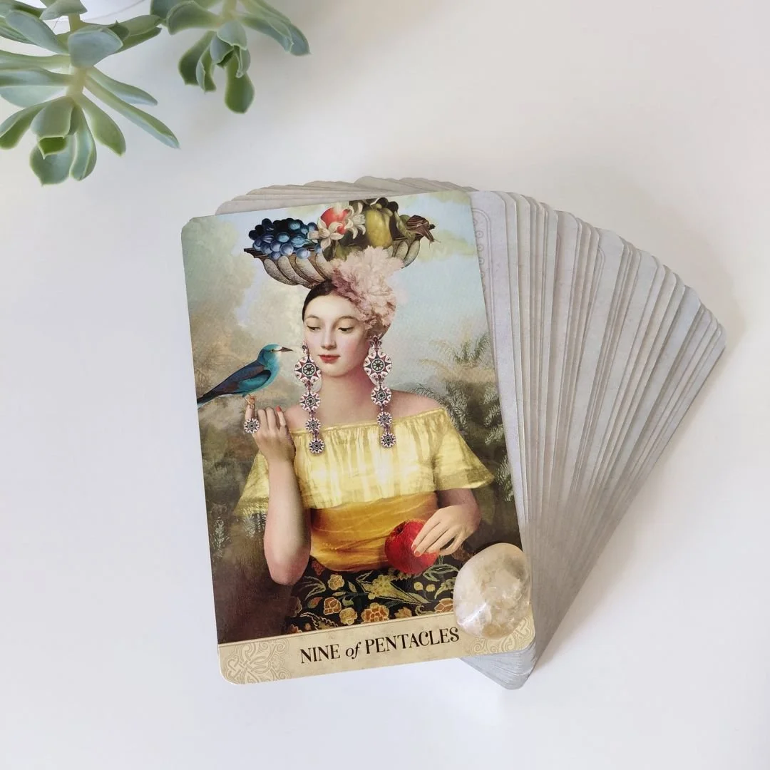 Flatlay of a fanned Tarot of Mystical Moments deck by Catrin Welz-Stein with the Nine of Pentacles card face up, topped with a polished yellow citrine crystal near succulent greenery.