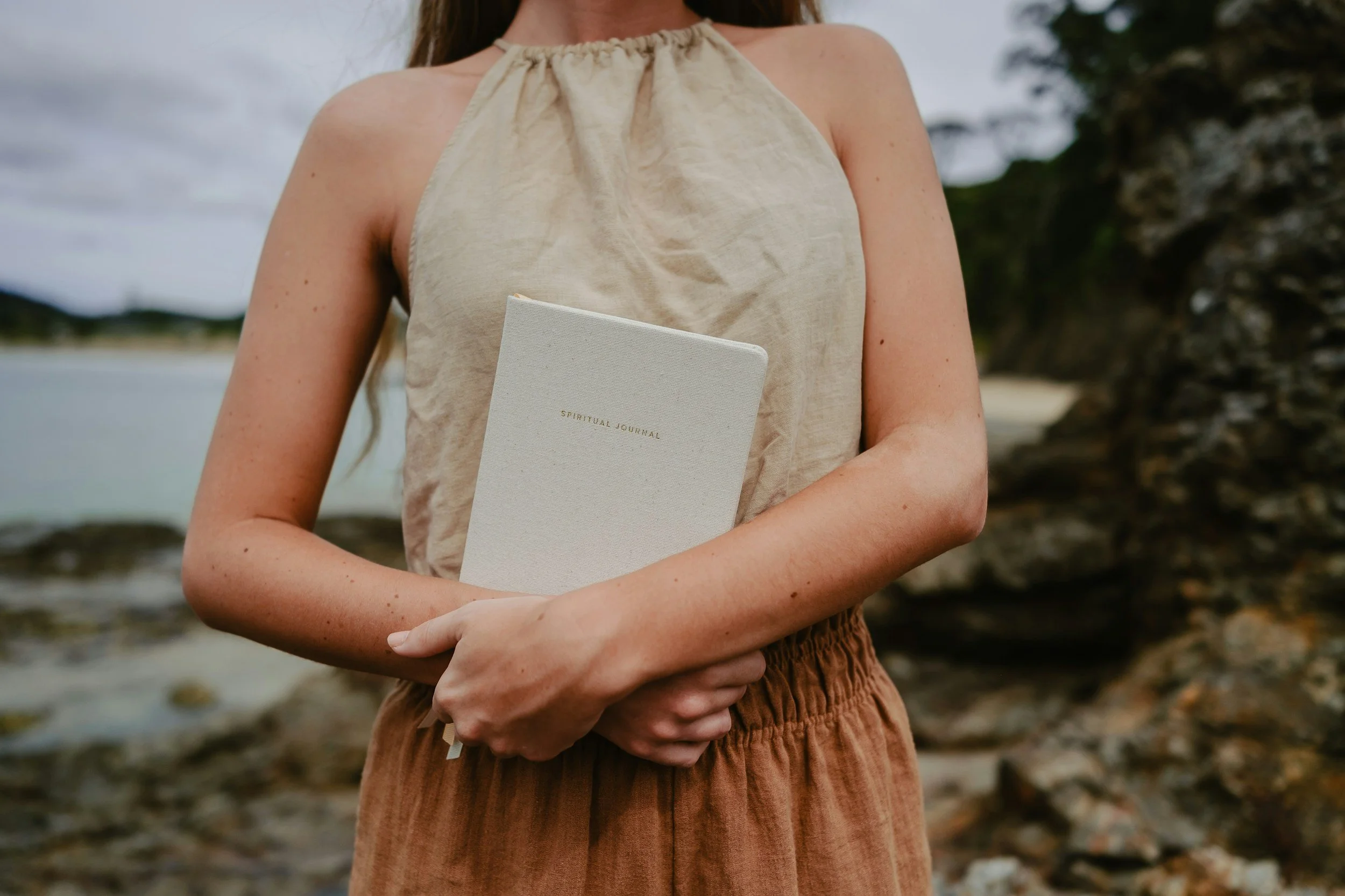 Grounded woman in linen clothing holding a spiritual journal outdoors by a calm river shore.