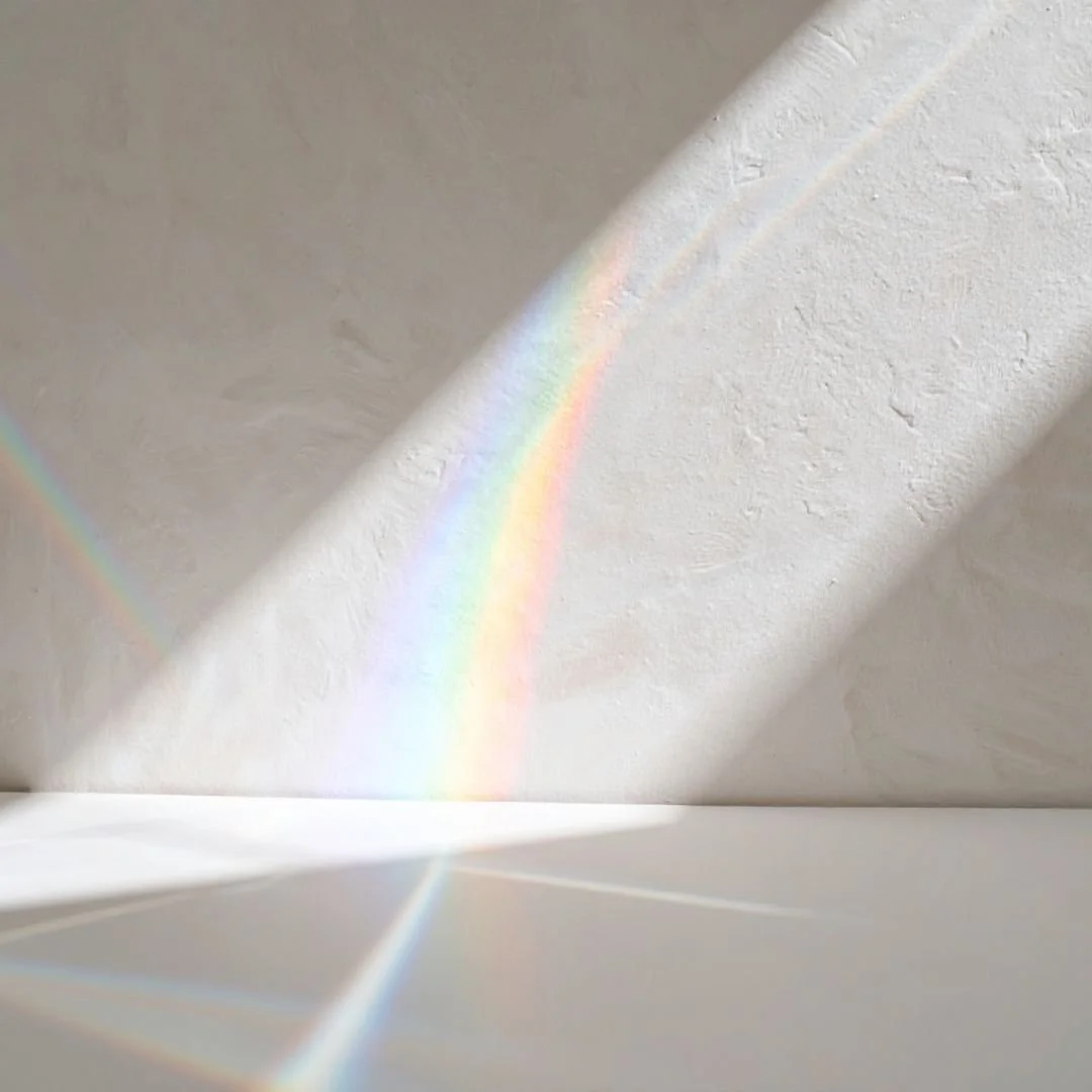 Rainbow light refraction from a crystal prism cast across a cream textured wall and smooth white tabletop, symbolizing distance energy healing for a virtual Reiki Level 2 training course.