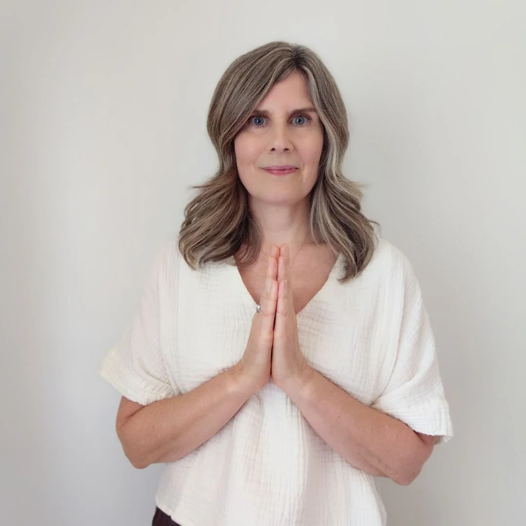 Claire, a spiritual alignment practitioner, standing in a gassho prayer position with her hands at her heart chakra against a pale cream wall, wearing an organic cotton blouse.