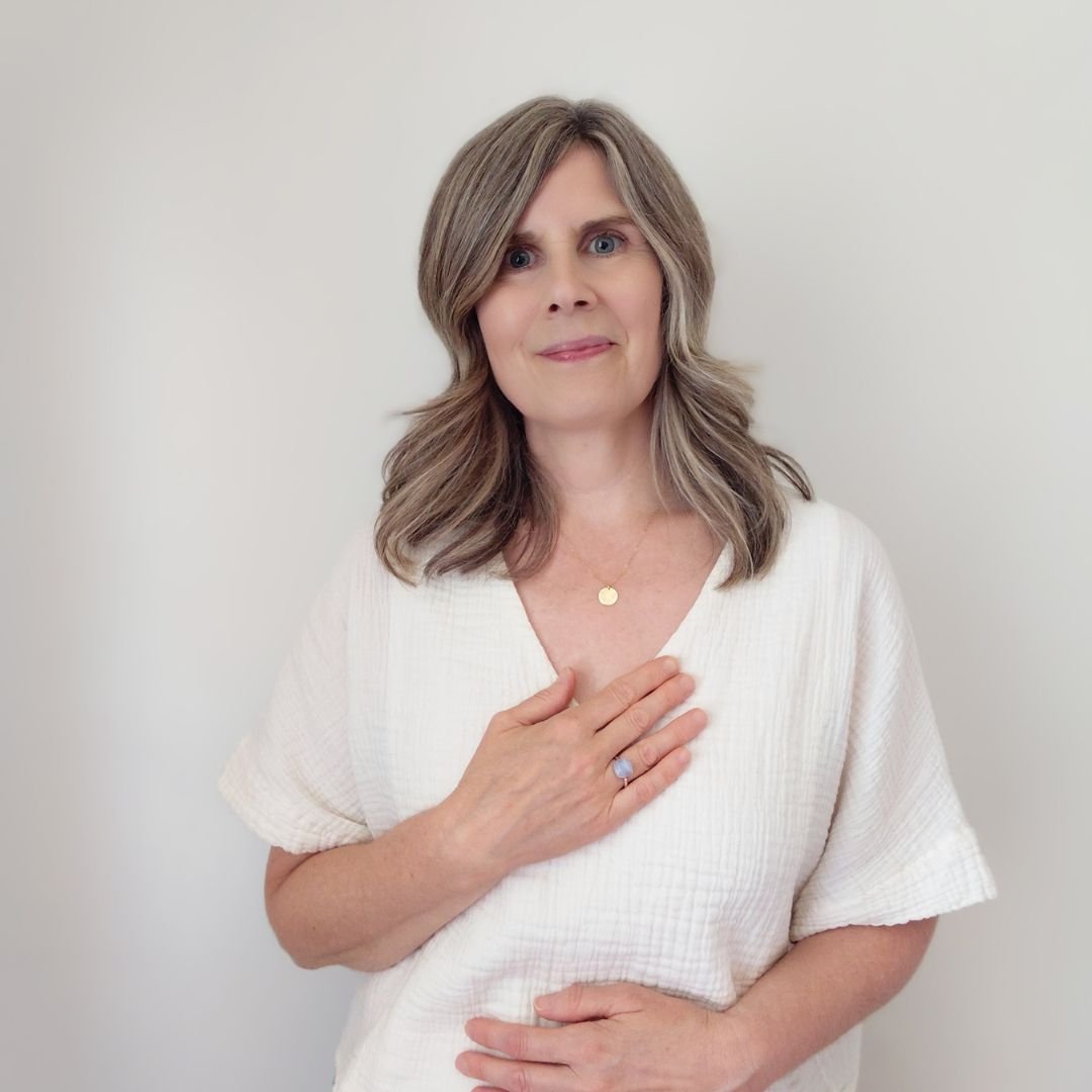 Spiritual alignment mentor Claire with one hand on her heart chakra and the other on her sacral chakra, posing against a soft cream background for a healing services page.