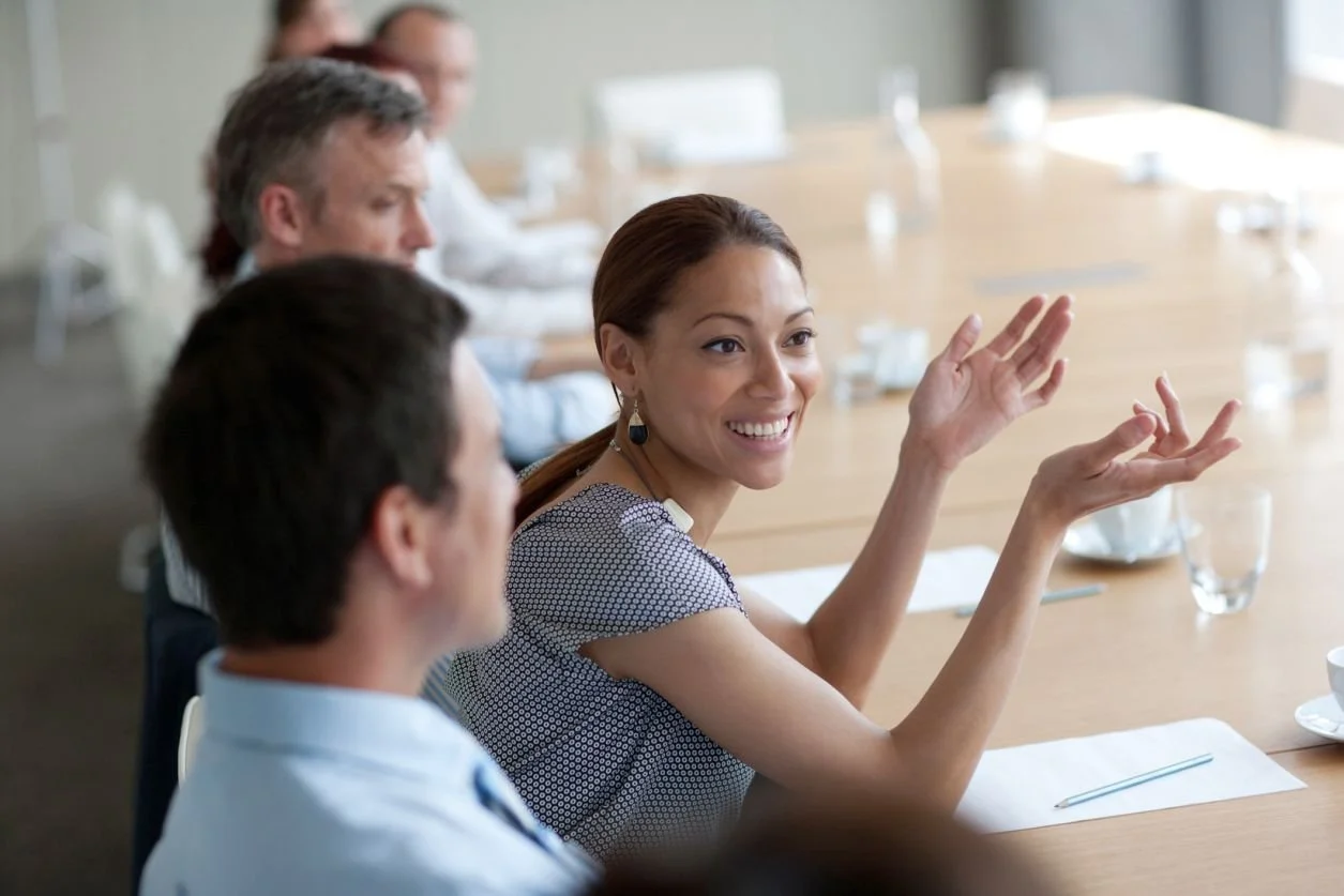 A woman is smiling and gesturing as she speaks during a meeting at a conference table. Several other people are seated around the table, listening attentively.