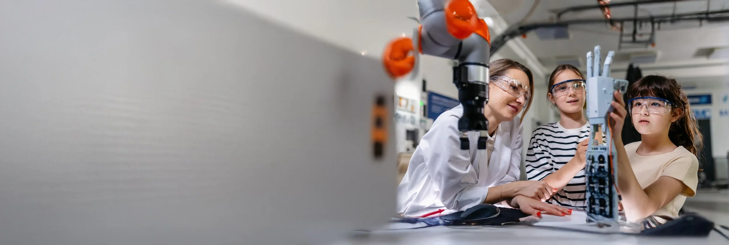A woman and two young girls in safety goggles working with robotic equipment in a laboratory setting.
