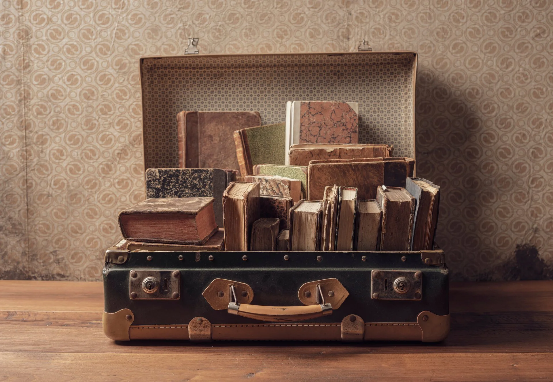 Open vintage suitcase filled with worn, antique books placed on a wooden surface with a patterned wallpaper background.